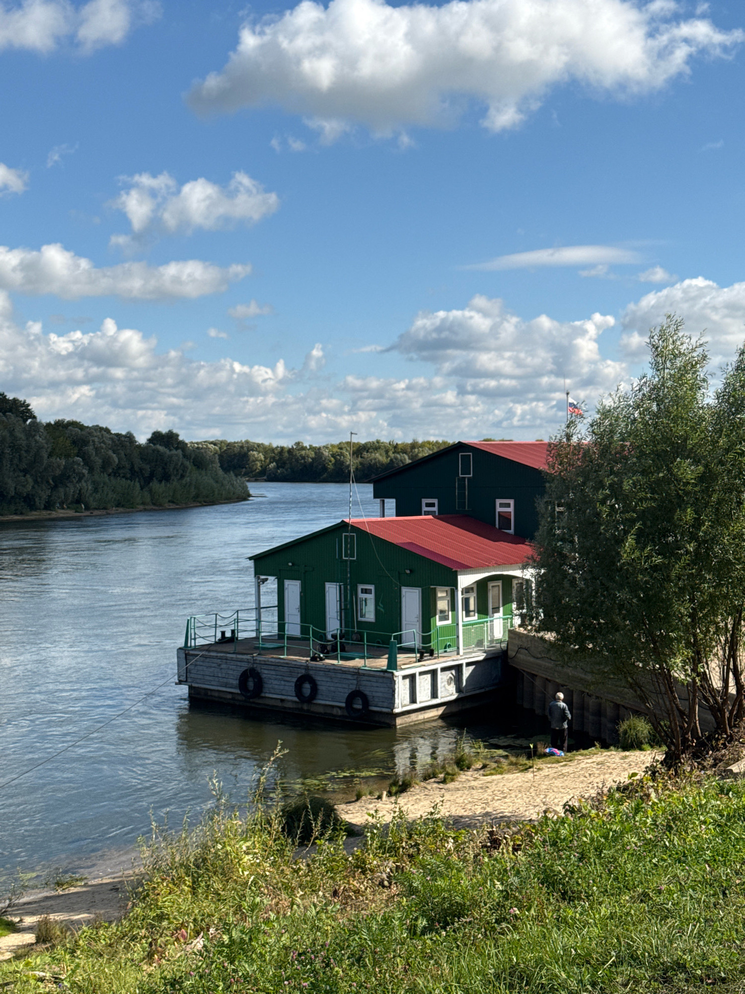 A wide, calm river flows on the left side of the image, with a lush green forest lining the opposite bank. In the foreground, a multi-level green building with red roofs is situated on a floating platform on the river. The platform has black tires acting as fenders along its side. A Russian flag flies from a pole atop one of the buildings. On the sandy riverbank to the right of the floating structure, a person stands facing away from the camera, looking towards the water. The area is covered with green grass and some smaller plants. The sky is blue with scattered white clouds. The image captures a serene riverside scene with a unique floating architectural feature. This is likely a location along the Oka River in Ryazan, Russia.