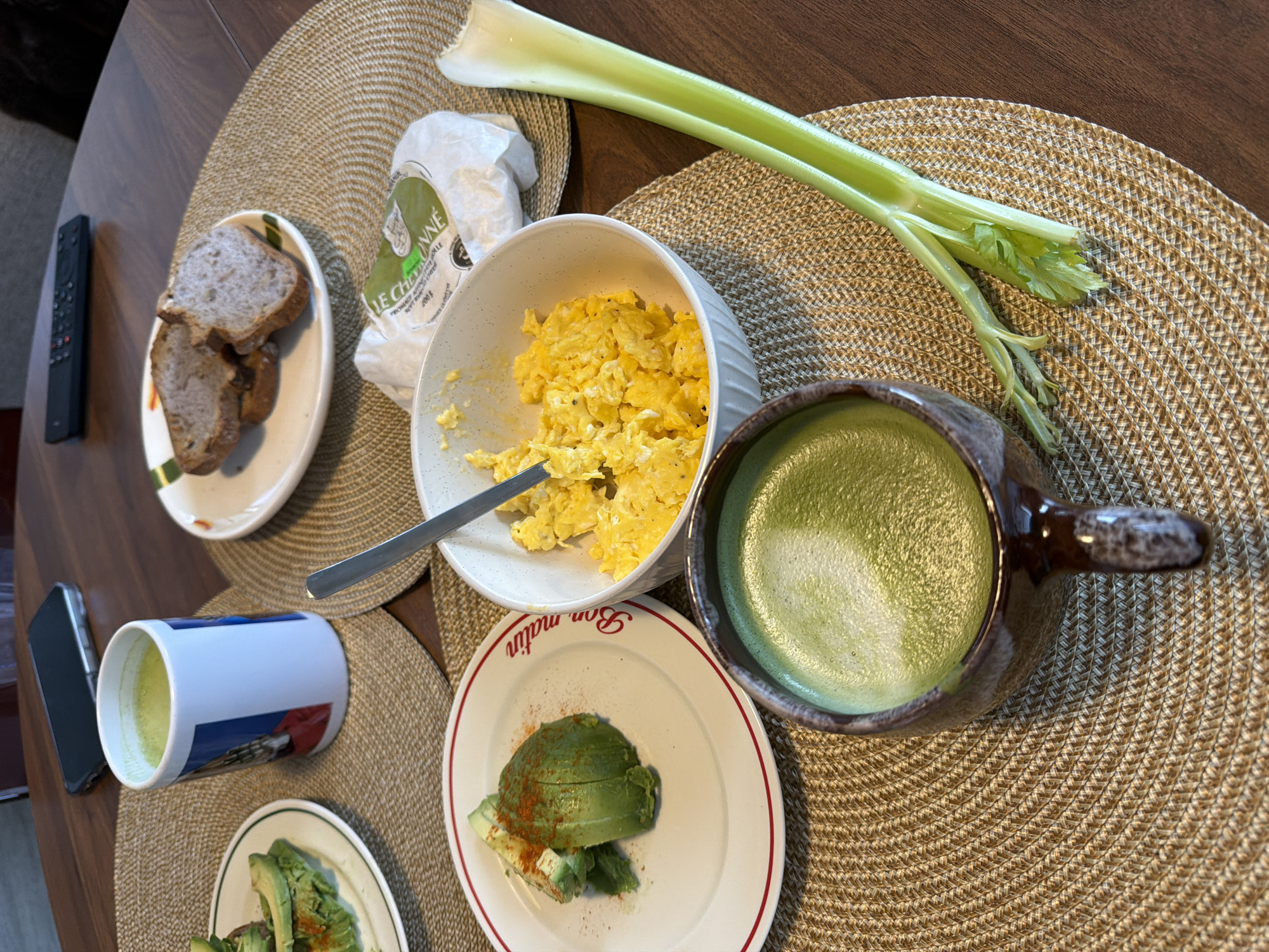 On a brown, textured placemat, a breakfast spread is arranged. The meal consists of scrambled eggs in a white bowl with a silver spoon, sliced avocado on two plates with red trim, a piece of celery, and slices of bread on a white plate with green trim. A package of "Le Chevrionne" cheese is near the bread. Two mugs containing a green beverage are also present. One mug is white with a blue design, and the other is a brown, stoneware-style mug. A smartphone and a black remote control are also on the table. A section of a dark brown wooden table is visible. The words "Bon Matin" are seen on one of the avocado plates.
There's no specific information in the image to pinpoint the precise location of the breakfast setting within the city of Montreal, Canada.
