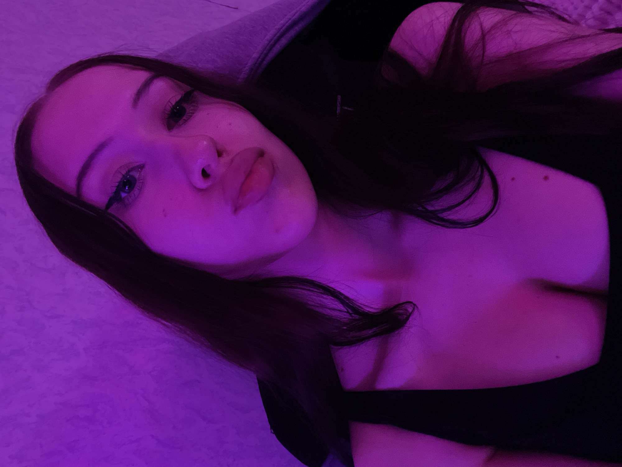 The image is a close-up, front-facing selfie of a young woman with long, dark hair, taken in a dimly lit environment. She is wearing a black top that reveals her décolletage. The scene is illuminated by a strong purple light, casting a distinct color cast over everything. Her makeup includes winged eyeliner. She appears to be indoors, with a textured wall visible in the background. There is no information available to place this image in Votkinsk, Russia.