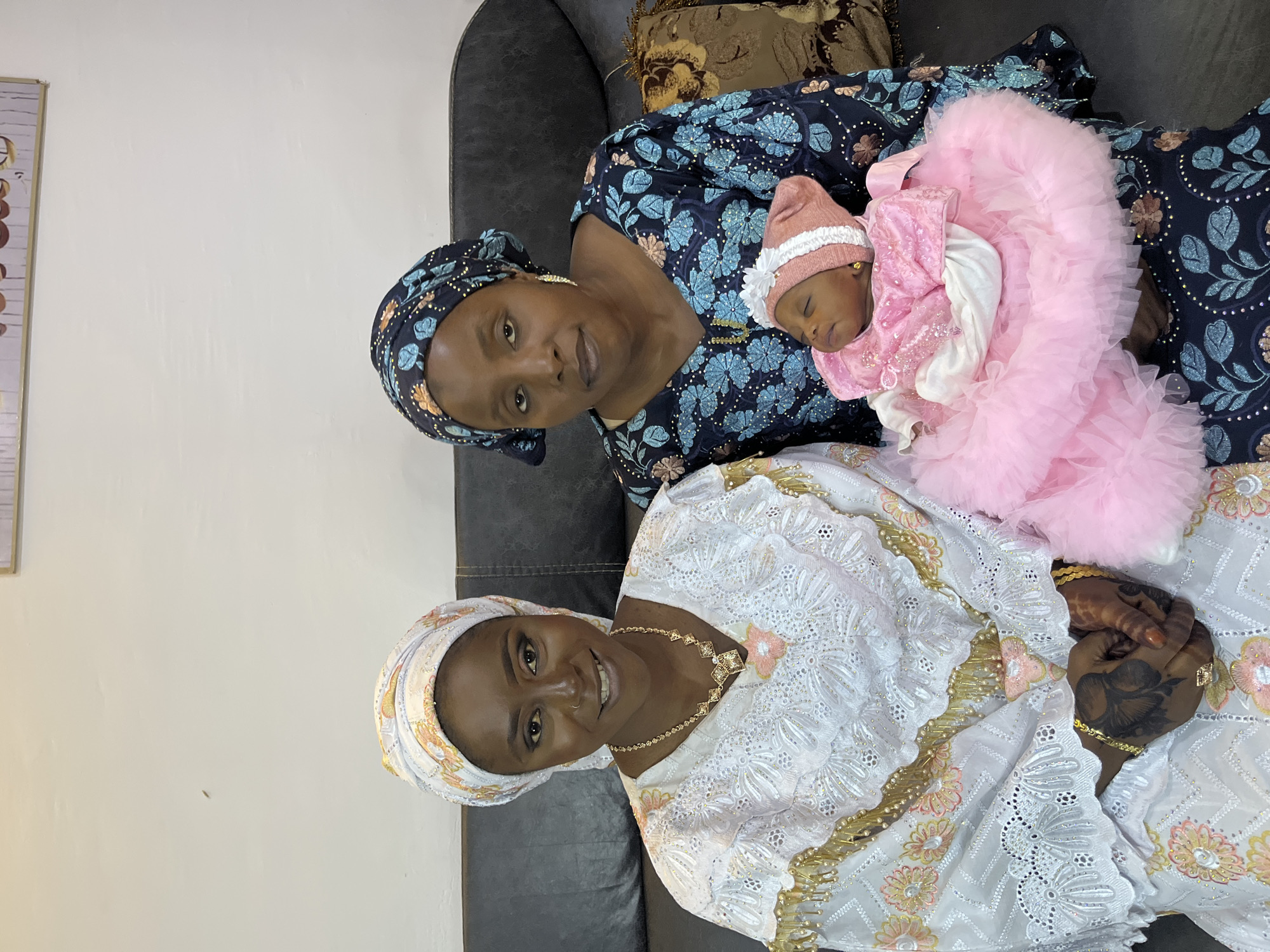 Three individuals, two adult women and an infant, are seated on a dark-colored couch, possibly in a residential setting in Jos, Nigeria. The woman on the left is wearing a white and gold traditional dress and a patterned headscarf. She is smiling and looking at the camera. The woman on the right is wearing a dark blue and floral print dress and a matching headscarf. She is holding a sleeping infant dressed in a pink outfit and bonnet. The infant is positioned in the crook of her arm, facing slightly away from the camera. The environment appears to be indoors, with a plain wall behind the subjects. A framed artwork is visible on the wall above them. The lighting is even, suggesting a well-lit indoor space. The overall scene depicts a family gathering or celebration.