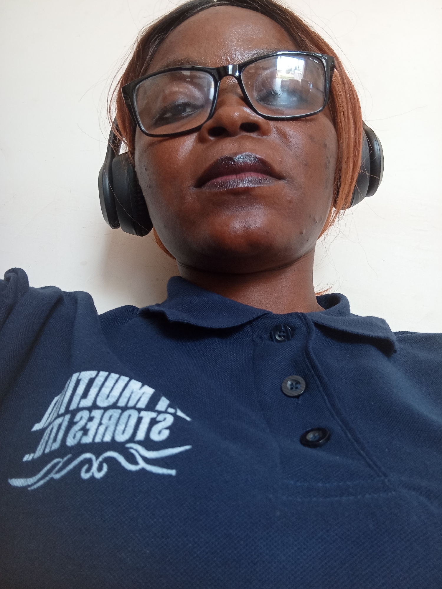 A medium shot shows a woman wearing headphones, glasses, and a navy blue collared shirt with a logo. The woman is looking directly at the camera. The logo on her shirt appears to be white lettering that reads "MULTUM STORES IN". The background is a plain white wall.
