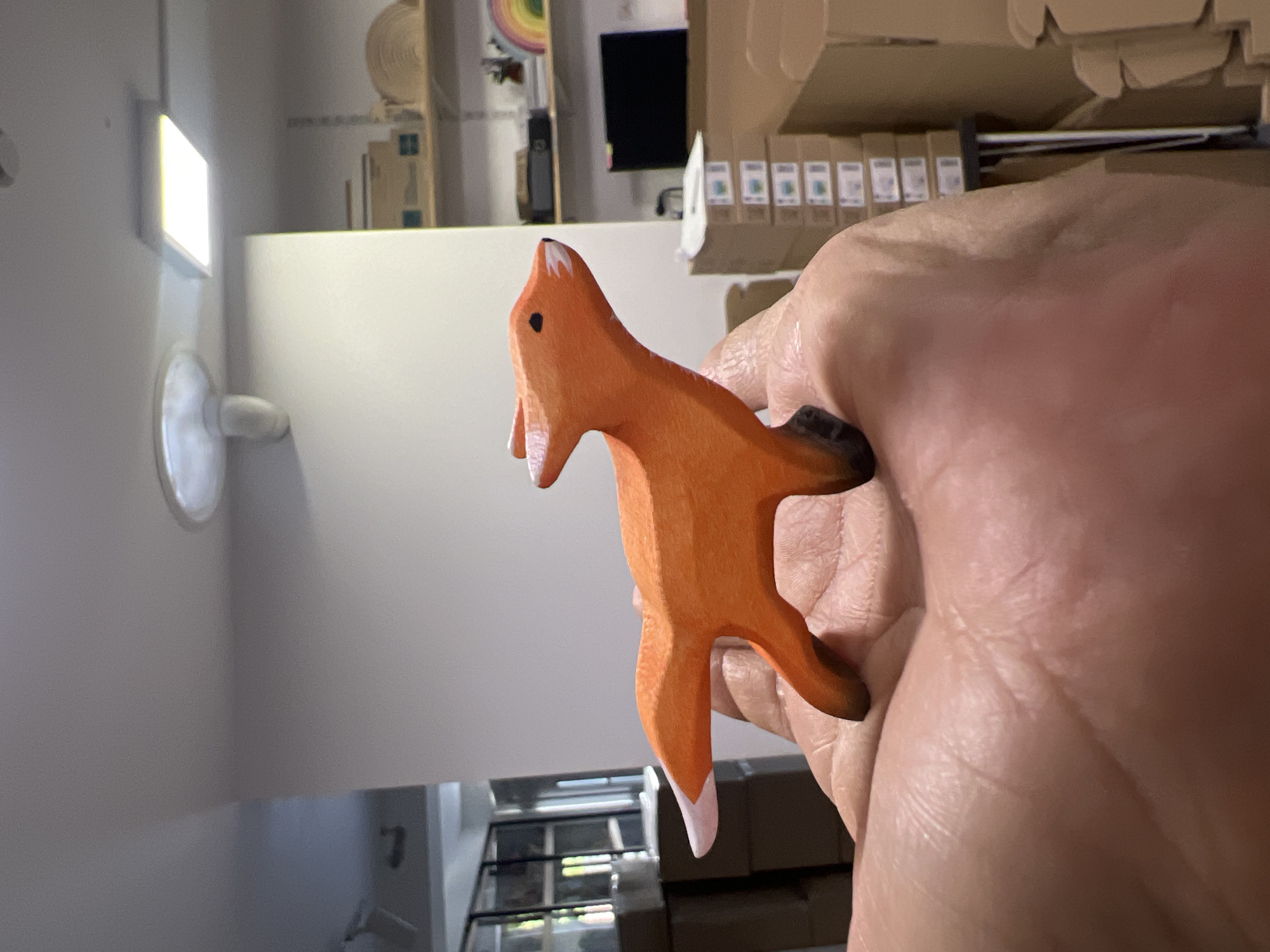 A person is holding a small, orange, wooden fox figurine in their right hand. The fox is depicted in profile, with its head tilted upwards and its mouth slightly open. It has black eyes and white tips on its ears and paws. The figurine is brightly lit, casting a subtle shadow on the hand.
The background is out of focus but suggests an indoor setting. To the left, a bright, rectangular light fixture is visible on a wall, and below it, a circular object, possibly a fan or vent. Further back, shelves are lined with what appear to be boxes. Some of these boxes have labels with small, colorful graphics.
The overall impression is that of a casual snapshot, likely taken indoors during daylight hours, given the illumination from the fixture. The focus is clearly on the wooden fox and the hand holding it. There is no discernible text in the image.