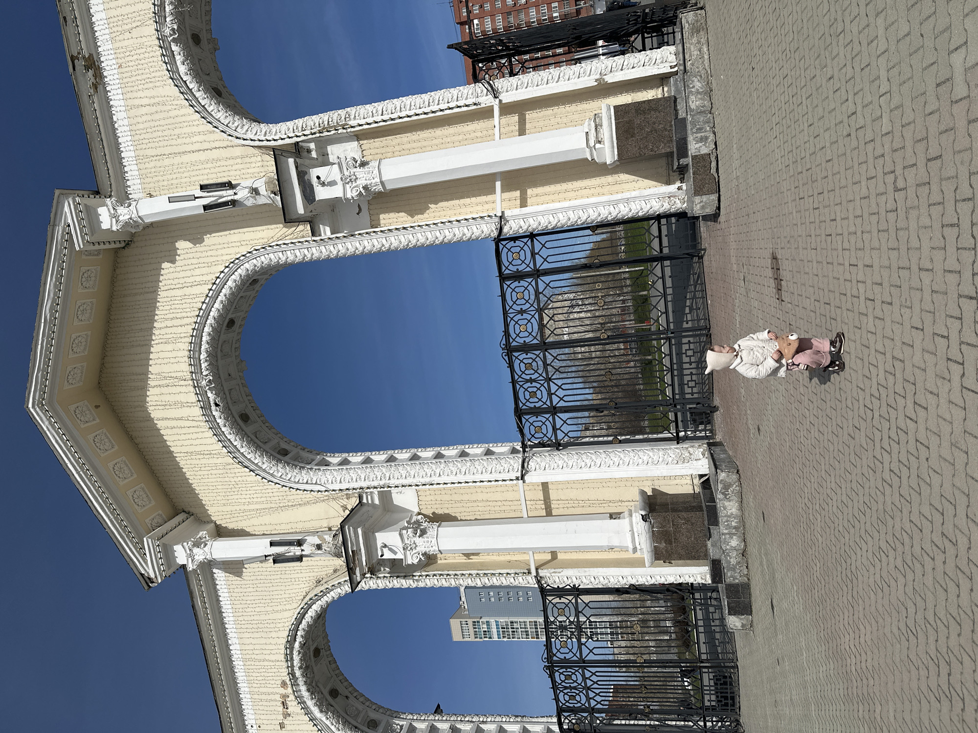 A low-angle, outdoor shot depicts a child walking on a brick-paved area framed by a light-colored, ornate archway structure, under a clear, blue sky. The archway structure features intricate architectural details and a black iron fence behind it. The child is wearing a white jacket, hat, and pink pants, and is holding a light-colored toy or object. A modern building can be seen through one of the archways. Based on the architectural style and design, the location appears to be in the city of Yekaterinburg, Russia.