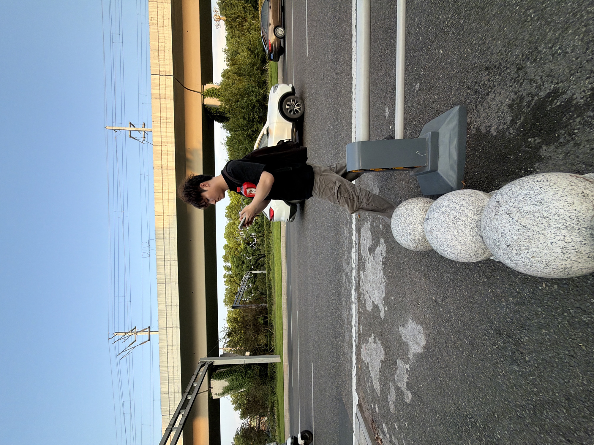 The image depicts a street scene in the city of Jiangjin, China. A young East Asian male, wearing a black t-shirt, grey cargo pants, and a black backpack, is walking on a sidewalk. He is looking down at his phone, with a red bottle attached to his shirt by a strap. In the background, traffic moves along a multi-lane road, with a white SUV and a brown sedan visible. An elevated structure, possibly a bridge or overpass with overhead power lines, spans the background. Lush green trees line the side of the road. The foreground shows a section of asphalt pavement with a white line, and decorative granite spheres are lined up next to a grey barrier. The sky is clear and blue, indicating it is daytime.