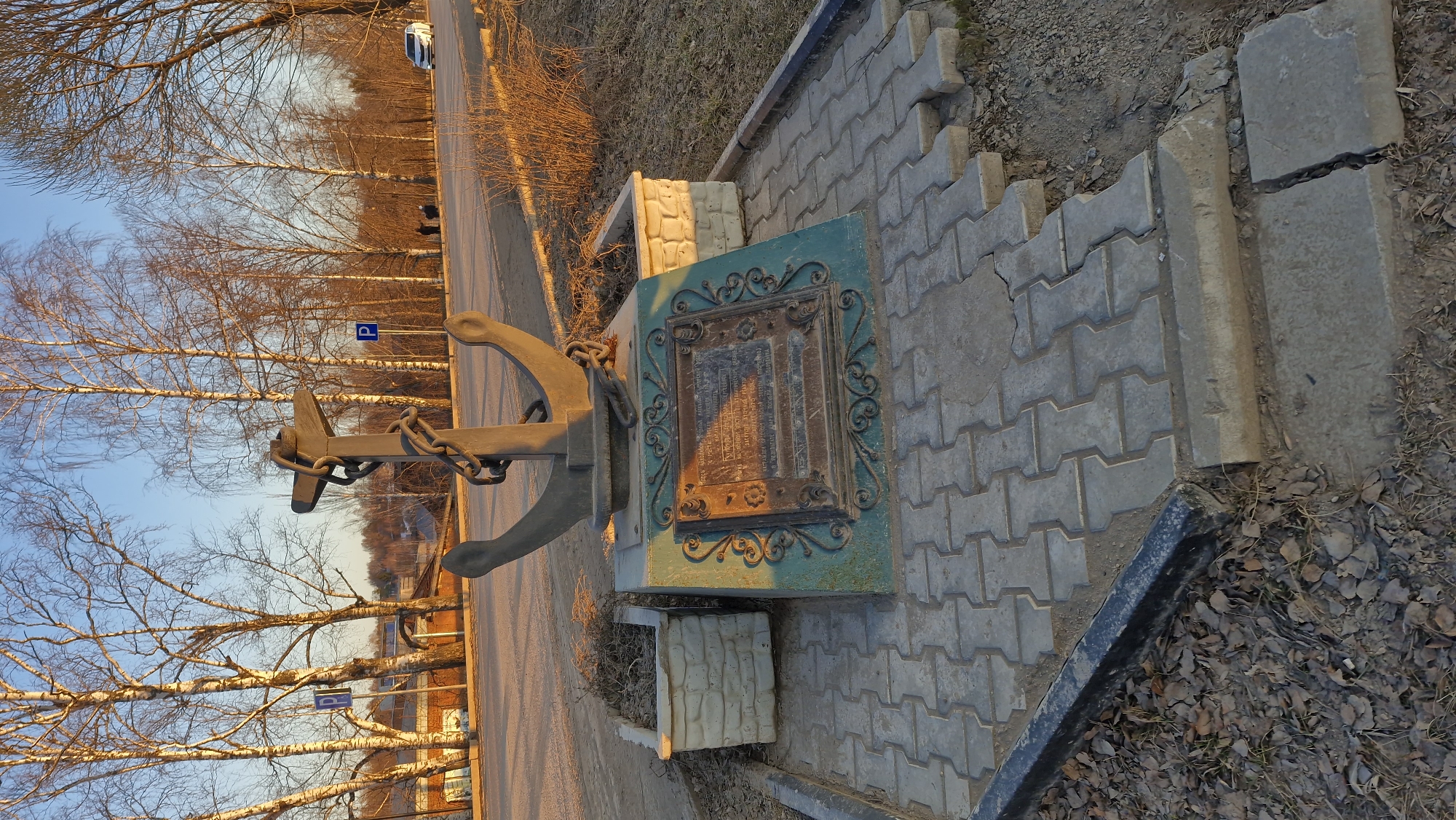 The image shows a monument featuring an anchor and a plaque in Otchor, Russia. The anchor, made of dark metal, is prominently displayed, connected to a plaque encased in a decorative green frame. The plaque appears to have an inscription. The monument is positioned on a paved area constructed with interlocking gray bricks, and is flanked by other concrete blocks or structures. The setting is outdoors, adjacent to a road lined with bare trees suggesting either late autumn or early spring. The sky is visible, with a gradient of blue to a soft sunset hue. There is a parking sign and a car visible in the background along the road. The ground around the paved area is covered with dry leaves.