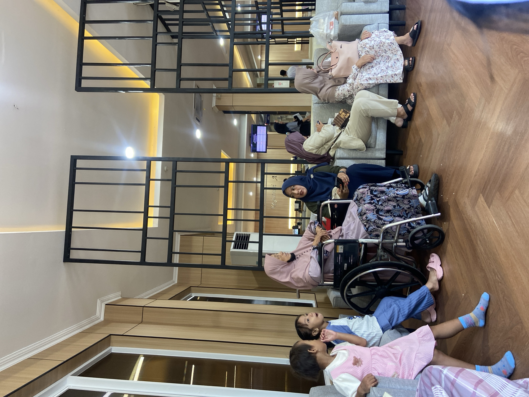 The image shows a waiting area, likely in a hospital or clinic, in Sidoarjo, Indonesia. There are several people present. Three women are seated on a grey sofa, two are wearing hijabs and floral dresses. Two people are in wheelchairs. Two children are lying on the floor in front of the wheelchairs. The interior features a wooden floor, a divider with a black metal frame, and recessed lighting.