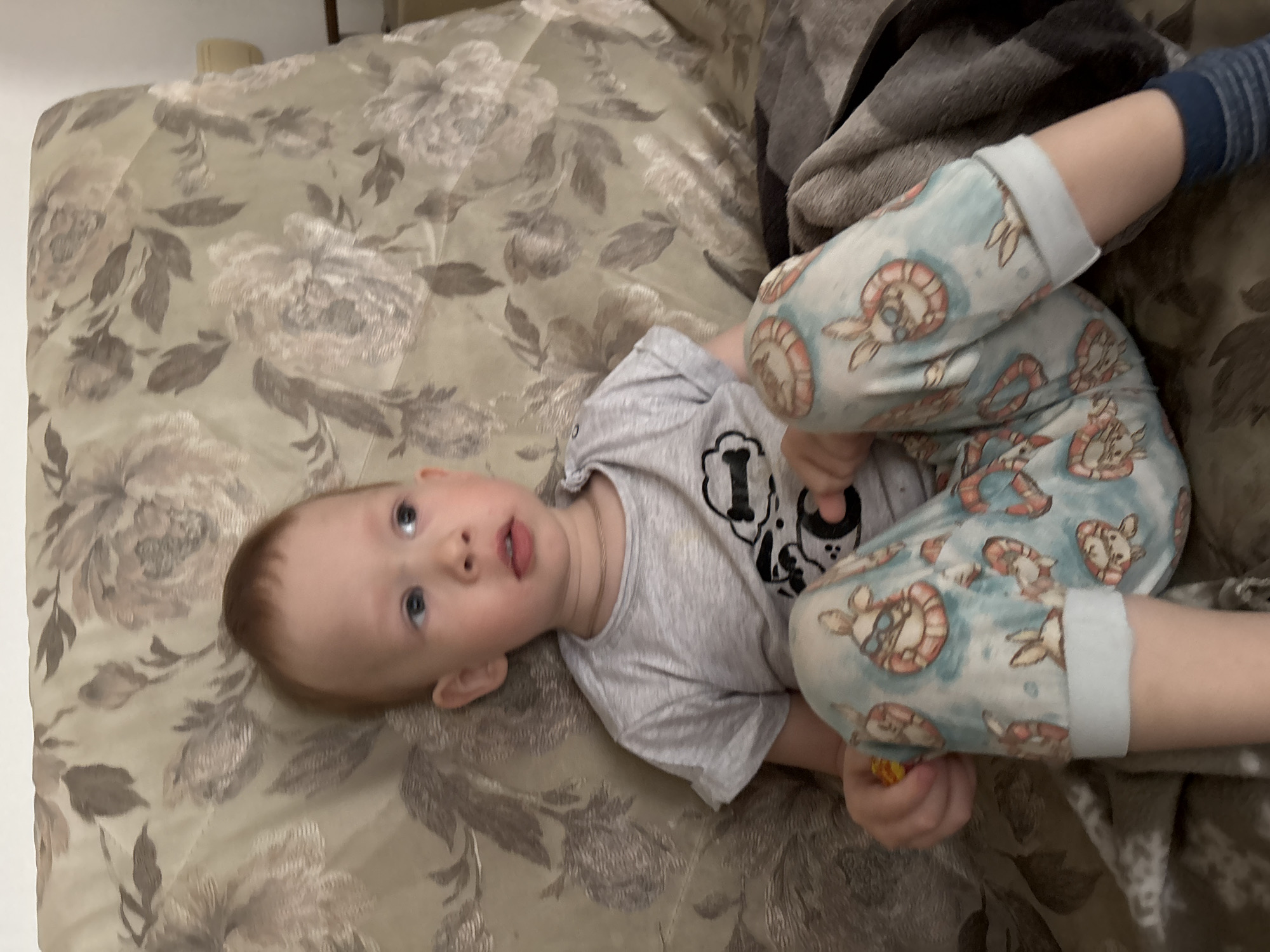 A young child lies on a floral patterned couch, covered partially by a brown blanket. The child wears a grey shirt with a black and white design and light blue pants with cartoon rabbits. The child's right foot is clad in a blue striped sock. The child looks upwards. The environment is domestic, interior. The location is Ferma, Russia.