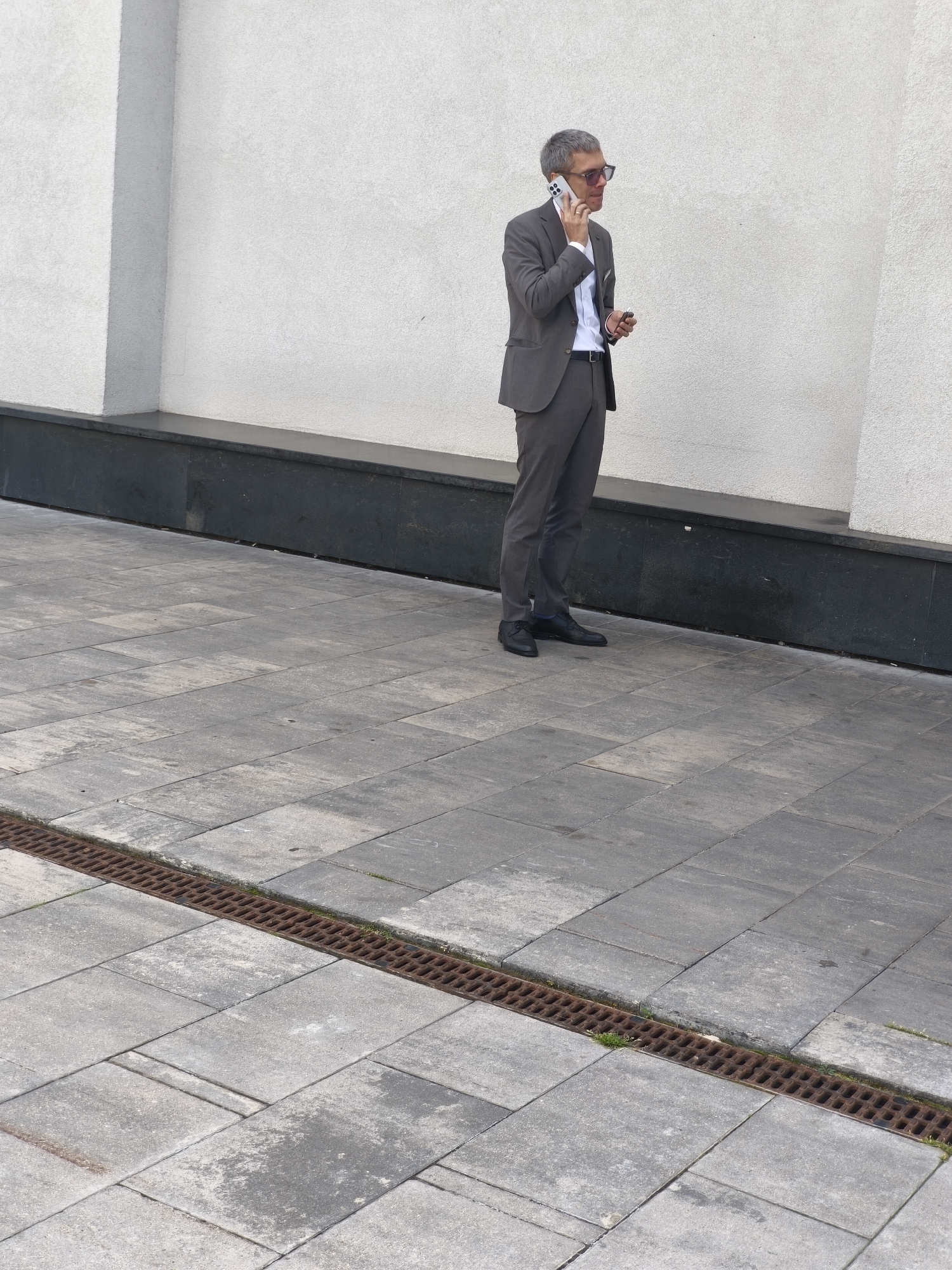 A man in a grey suit, white shirt, sunglasses, and black shoes is standing on a paved sidewalk and talking on a cell phone. He is holding a small object in his right hand. The background is a textured white wall with a dark grey base. A metal grate runs along the edge of the sidewalk. The scene appears to be outdoors in an urban environment, possibly in Minsk, Belarus.