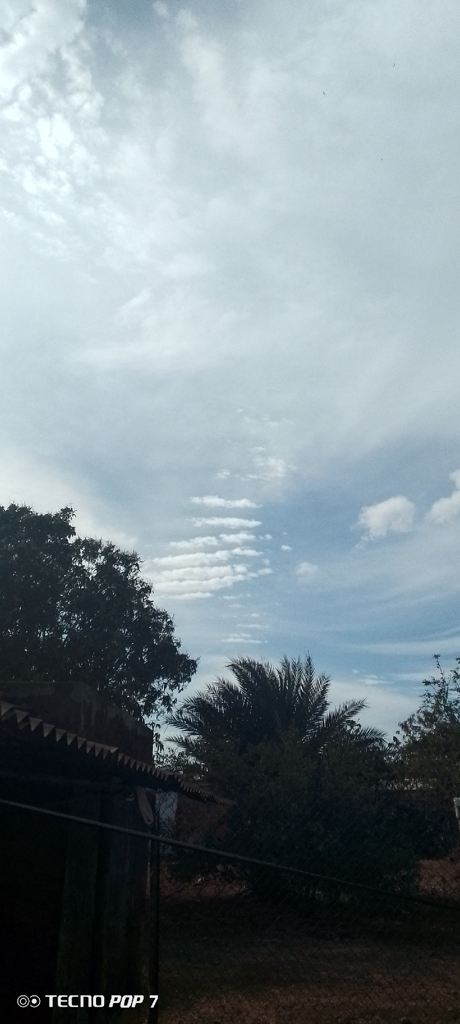 The image shows a partly cloudy sky above Altagracia, Venezuela. In the foreground, there are trees, a palm tree, and the corner of a building or structure with a zig-zag roofline. A fence is partially visible in the bottom right corner. The sky has wispy clouds and patches of blue. The image was taken with a Tecno Pop 7 mobile phone.