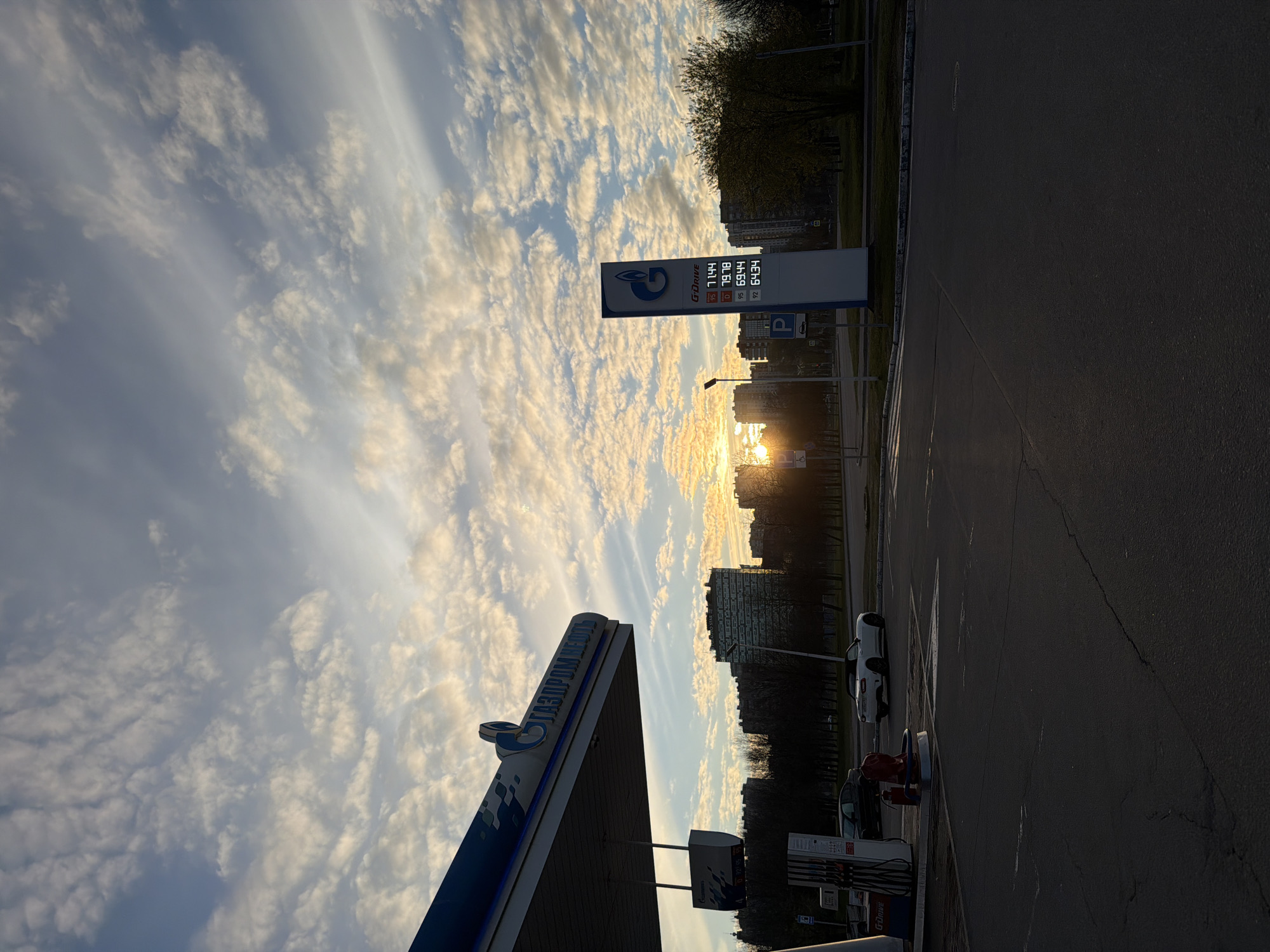 The image depicts a Gazprom gas station in Saint Petersburg, Russia, at dusk or dawn. The sky is filled with textured clouds and a visible sun. A price sign displays fuel costs for different grades. A white car is parked near the pumps. In the background are buildings partially obscured by trees.