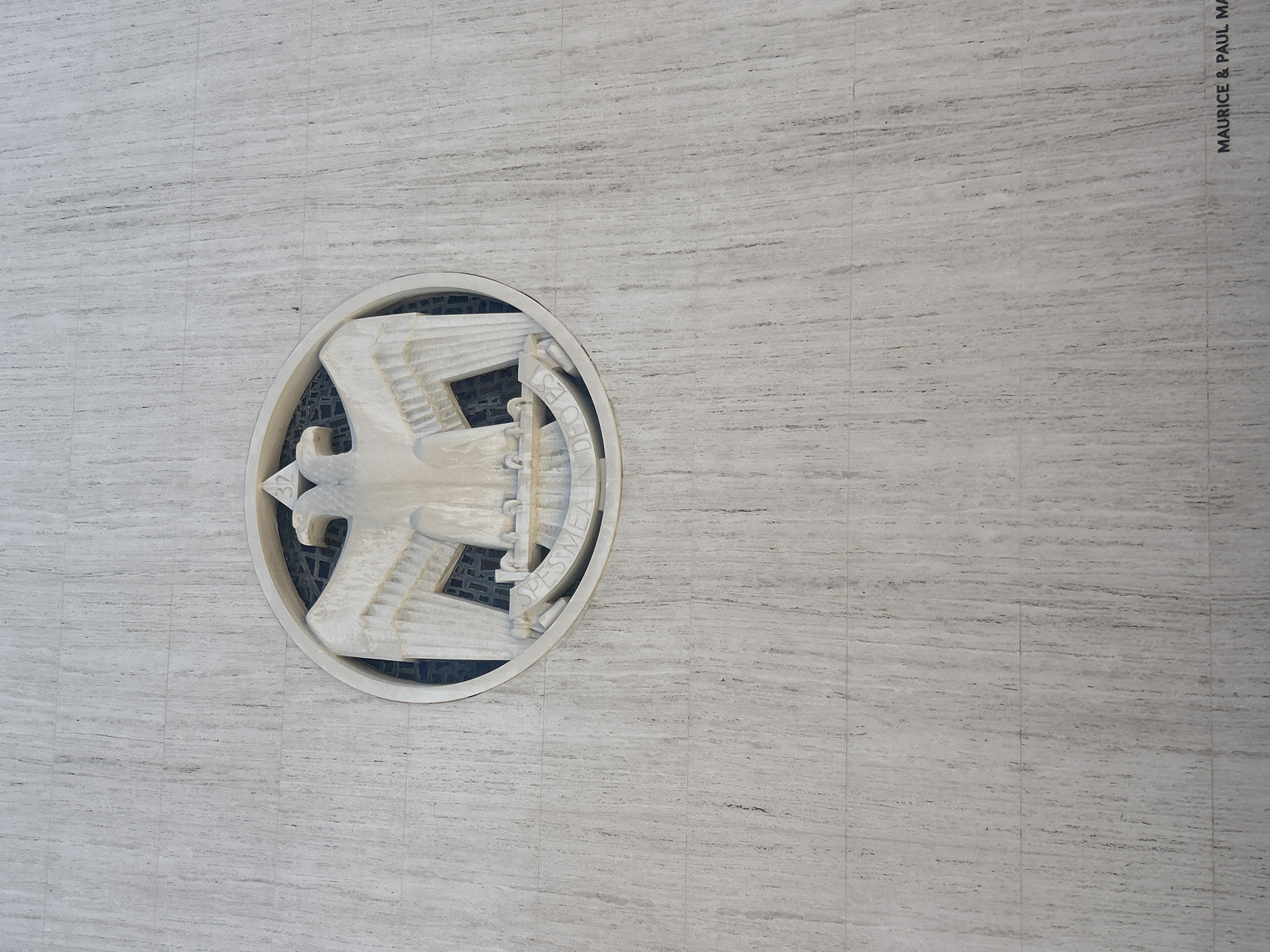 The image depicts a close-up view of a circular emblem affixed to a wall with a light-gray, striated texture. The emblem contains a stylized bird figure oriented towards the left, encircled by lettering. A dark material is visible behind the emblem. In the upper right corner, the text "MAURICE & PAUL MARC" is printed vertically. The emblem is located on the wall of the United States Court of Appeals for the Ninth Circuit located in the city of Los Angeles, United States of America.