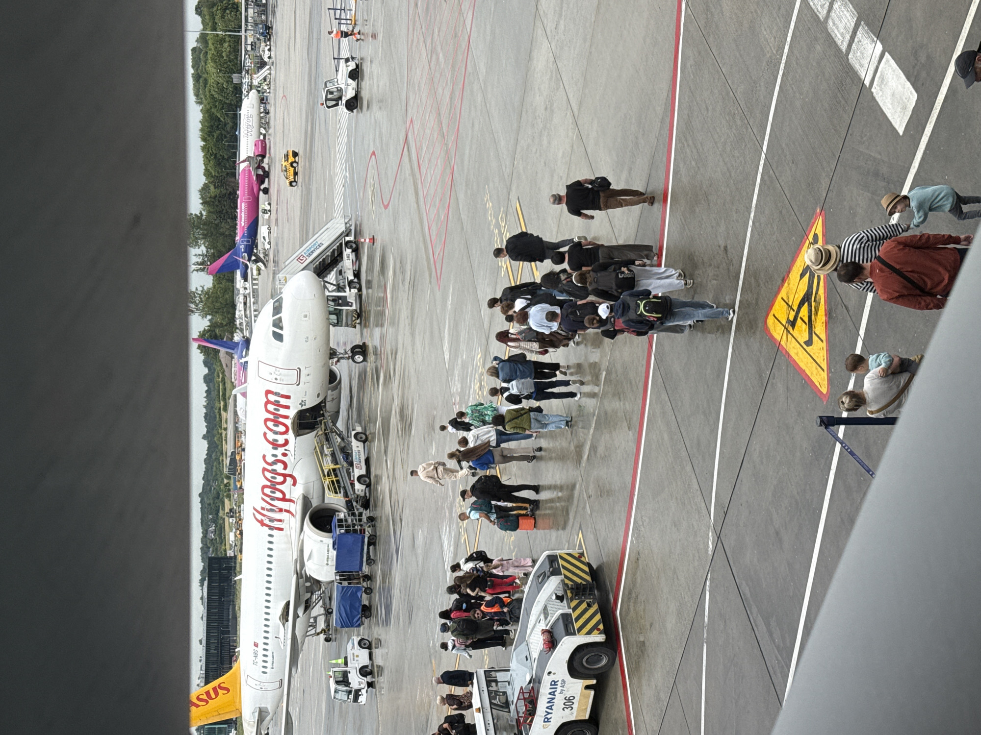 In Balice, Poland, at an airport tarmac, a group of passengers are boarding an airplane. The aircraft is white with orange and red accents and the logo "flypgs.com". A yellow and white tug vehicle is positioned near the front of the plane, with the text "RYANAIR by ASP 306" visible. Several other vehicles, including a baggage cart and a fire truck, are present on the wet tarmac. In the background, another airplane with a pink and purple livery is visible, as are airport buildings and trees. The sky is overcast and dark. The passengers, dressed in casual attire, are walking towards the airplane with luggage. Some are carrying bags, while others are pushing strollers. A yellow warning sign with a depiction of a person slipping is painted on the tarmac.