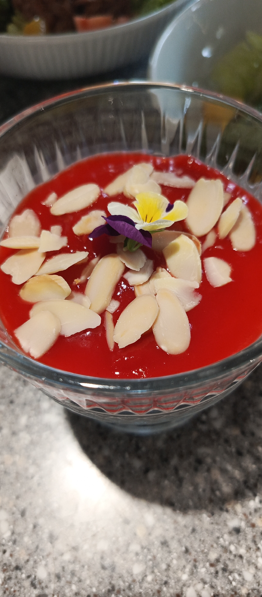A close-up shot shows a clear glass bowl filled with a vibrant red dessert. The dessert is topped with thinly sliced almonds and a small, delicate flower. The flower has yellow and white petals with purple accents, adding a touch of color and elegance to the dish. The red dessert has a smooth, glossy texture, suggesting it could be a fruit puree, mousse, or a similar preparation. The almonds are scattered generously, creating a pleasing visual contrast against the red background. The glass bowl has a decorative, faceted base, adding a subtle sparkle to the composition. In the background, slightly out of focus, are other dishes or bowls, hinting at a meal or gathering. The lighting in the image is soft, highlighting the textures and colors of the dessert. The overall impression is one of a beautifully presented, refreshing, and perhaps summery treat. There is no visible text in the image.