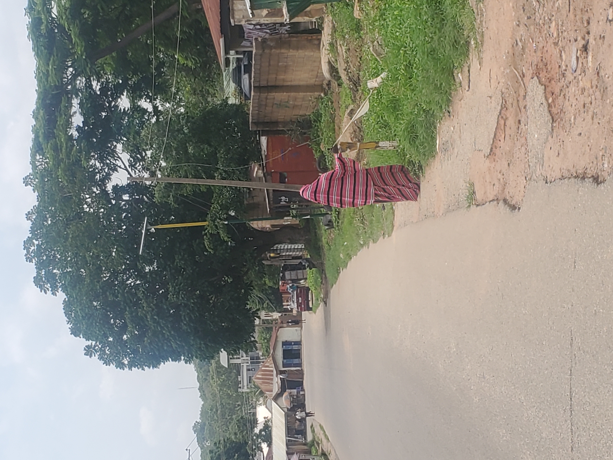 A person draped in a red and white striped garment stands on the side of a paved road, holding a white flag attached to a stick. The road is lined with various structures, including small shops with corrugated iron roofs, a red building, and a brick wall. Lush green vegetation grows alongside the road and around the buildings. In the background, trees and more buildings are visible, with a hazy sky overhead. The scene appears to be in a residential or commercial area of Jos, Nigeria.