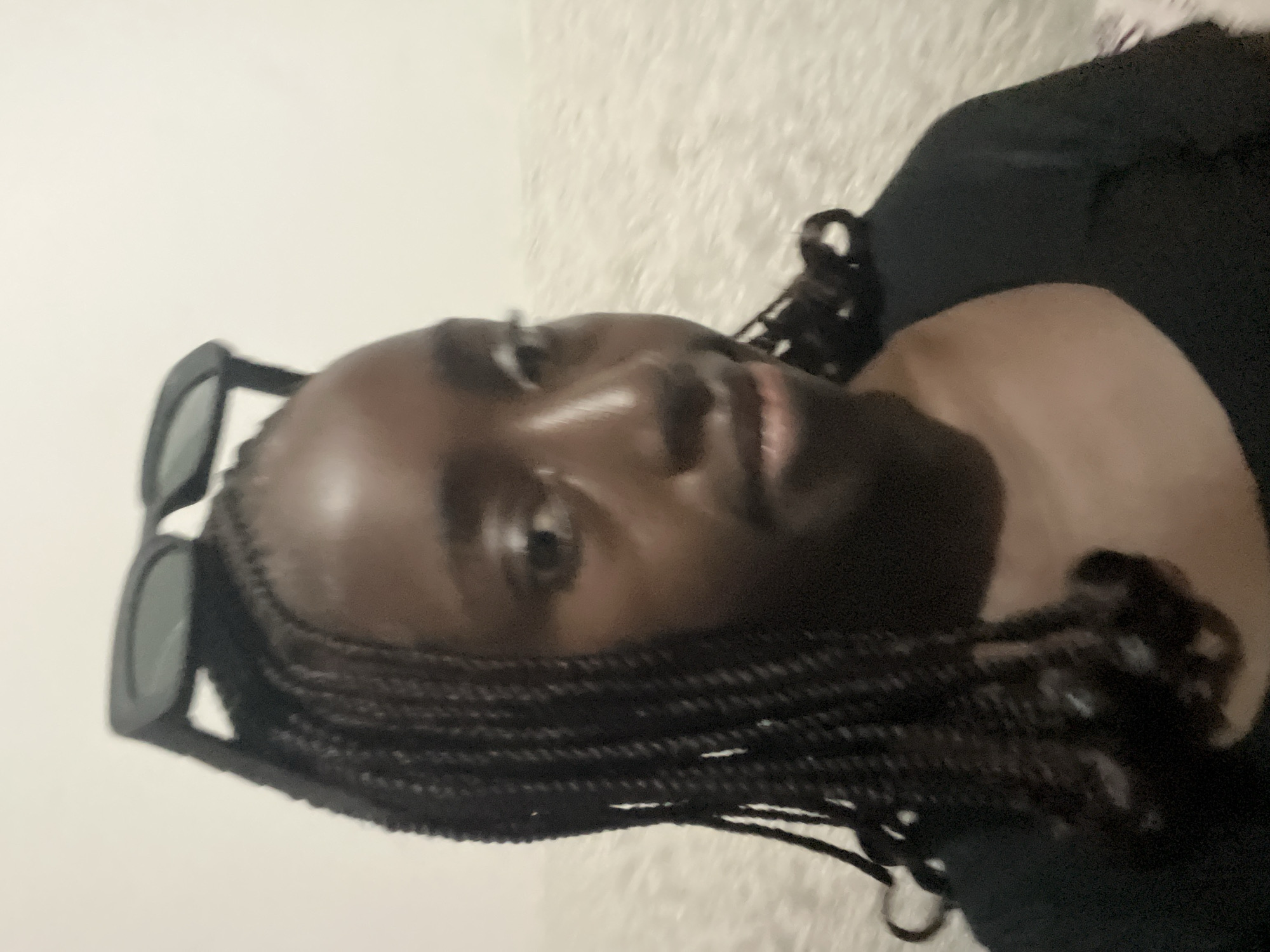 A dark-skinned woman with braided hair, sunglasses perched on her head, wearing a dark top is visible in the frame. The background appears to be a fuzzy white surface. No specific location in Abuja, Nigeria, can be determined from the image.