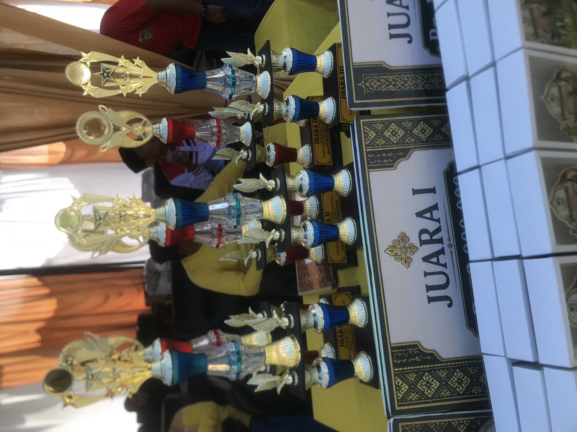 The image depicts an awards ceremony, likely held outdoors or in a covered area, with several tiers of trophies displayed prominently on a yellow tablecloth. The trophies are predominantly gold with blue and red accents, some featuring star and eagle motifs. In the background, people are visible, some wearing traditional clothing. Specifically, there are individuals in yellow and red shirts. The environment suggests a festive occasion, possibly a local competition or event. The presence of decorative curtains in the background and the arrangement of trophies indicate a structured award presentation. Although the exact location within Langkuta, Indonesia, cannot be determined solely from the visual information, the scene strongly suggests a community-focused event. The focus is on the awards, implying a celebration of achievement.