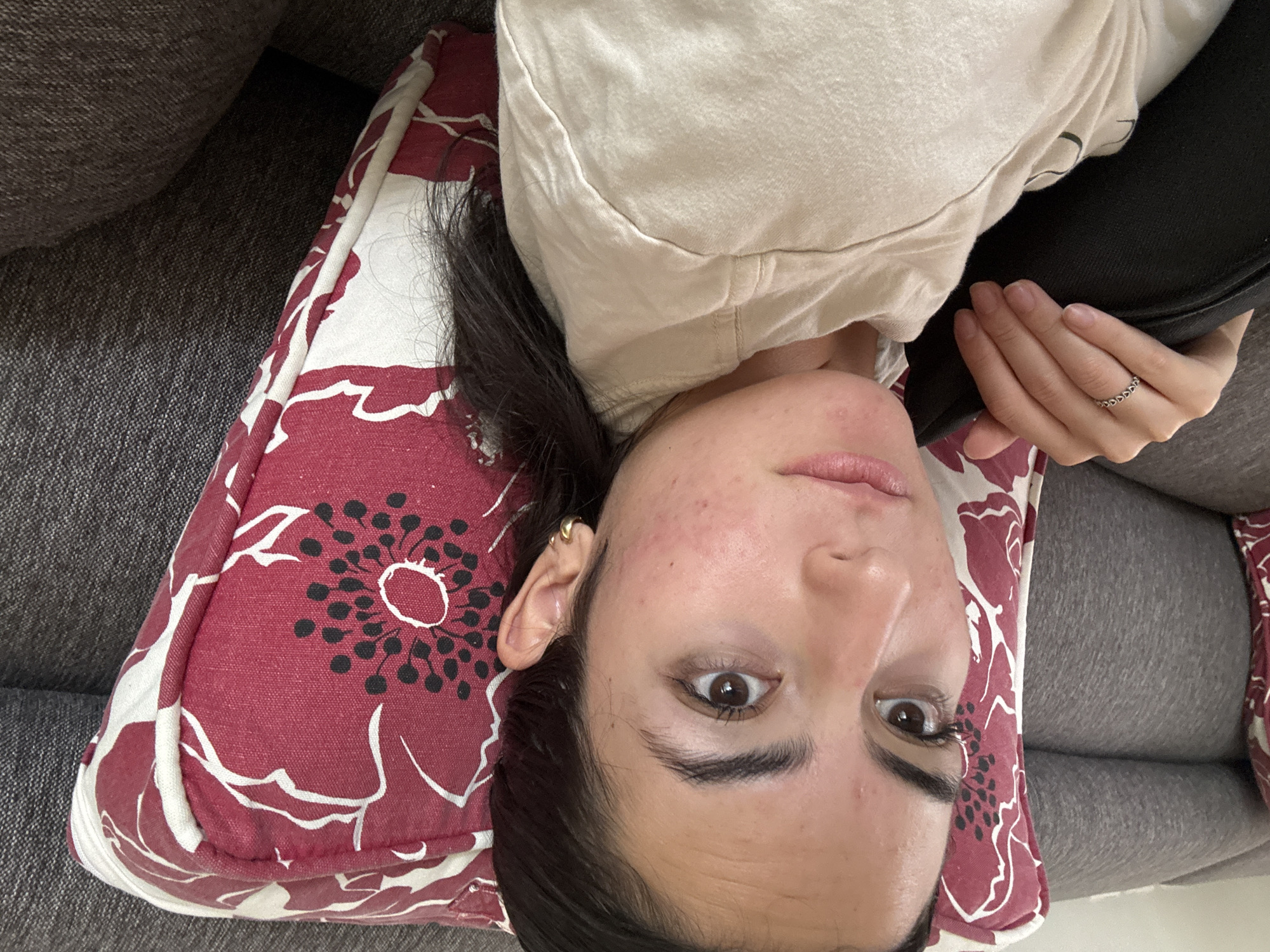 A woman is lying on a gray couch. She is resting her head on a red and white floral patterned pillow. She is wearing a light-colored hooded sweatshirt. She is holding a black object in her right hand, which has a silver ring on the ring finger. The woman's face is visible, and she is looking directly at the camera. The image was taken in an indoor setting. There is no information in the image to indicate that it was taken in San Bernardino Tlaxcalancingo, Mexico.