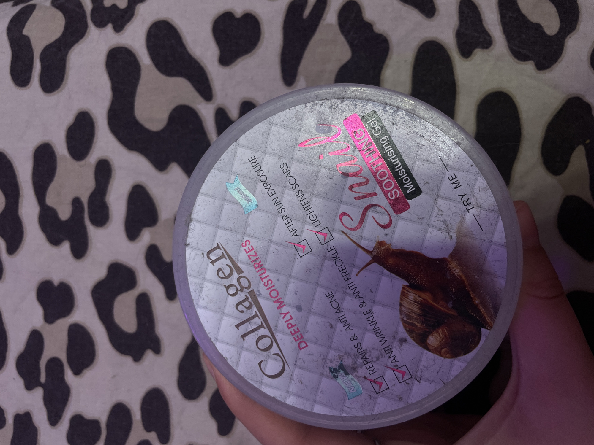 A person's hand is holding a round, clear plastic jar of "Collagen Deeply Moisturizes" and "Snail Soothing Moisturising Gel". The jar has a white lid with a textured surface and a holographic label. The label features a large image of a snail, the text "Collagen", "DEEPLY MOISTURIZES", "Snail SOOTHING", and "Moisturising Gel". There are also checkboxes next to claims like "REPAIRS & ANTI ACNE", "ANTI WRINKLE & ANTI FRECKLE", "AFTER SUN EXPOSURE", and "LIGHTENS SCARS". A small blue banner indicates "300ml". The jar is placed on a surface with a black and beige leopard print pattern. The background of the image is out of focus. There is no identifiable information about the location in Antonovka, Russia.