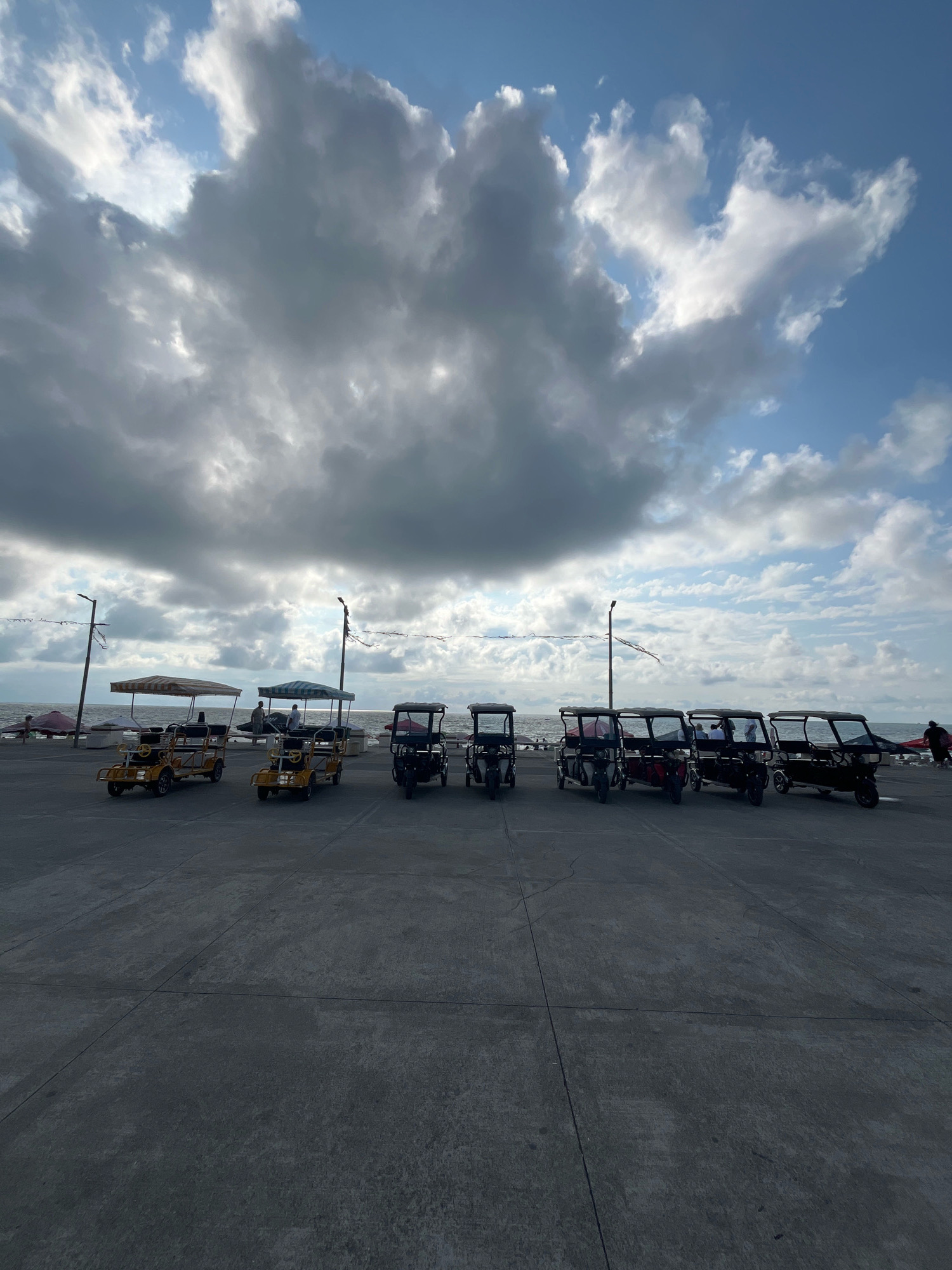 The image depicts a parking area near the Black Sea coast in Ureki, Georgia, under a sky filled with dramatic clouds. Several open-air, multi-passenger pedal carts are parked in a row on the paved ground. Two of these carts, yellow in color and with striped canopies, are positioned towards the left side of the frame. To their right, a line of darker colored, enclosed pedal carts extends across the scene, with some appearing to have passengers. In the background, the Black Sea is visible, with a faint horizon line. A few utility poles with lights and string decorations are scattered along the waterfront. Several individuals are visible, some standing near the carts and others further back on what appears to be a promenade or walkway. One person is seen carrying a red object near the far right of the frame. The overall atmosphere is overcast, suggesting a potentially breezy or transitioning weather condition.