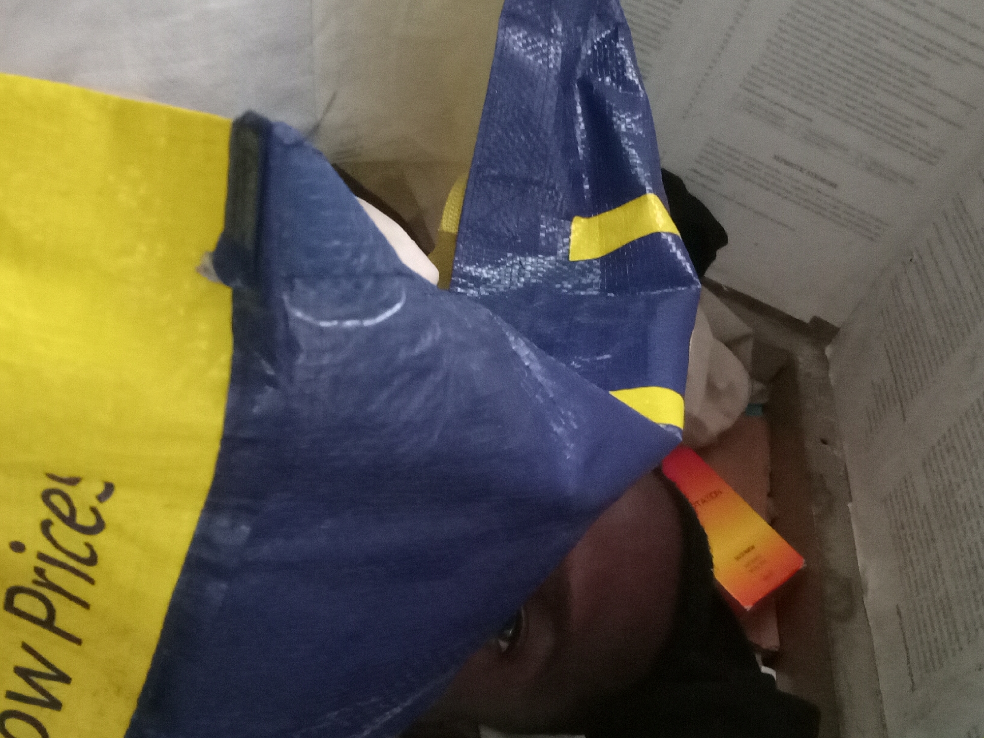 A person is partially visible inside a shopping bag with yellow and blue colors with the words "Low Prices" printed on the yellow part. The bag is located in Akwete, Nigeria. Other items appear to be inside the bag including what appears to be a cosmetics box. The inside of the bag shows a textured surface, possibly paper, where the contents are stored.