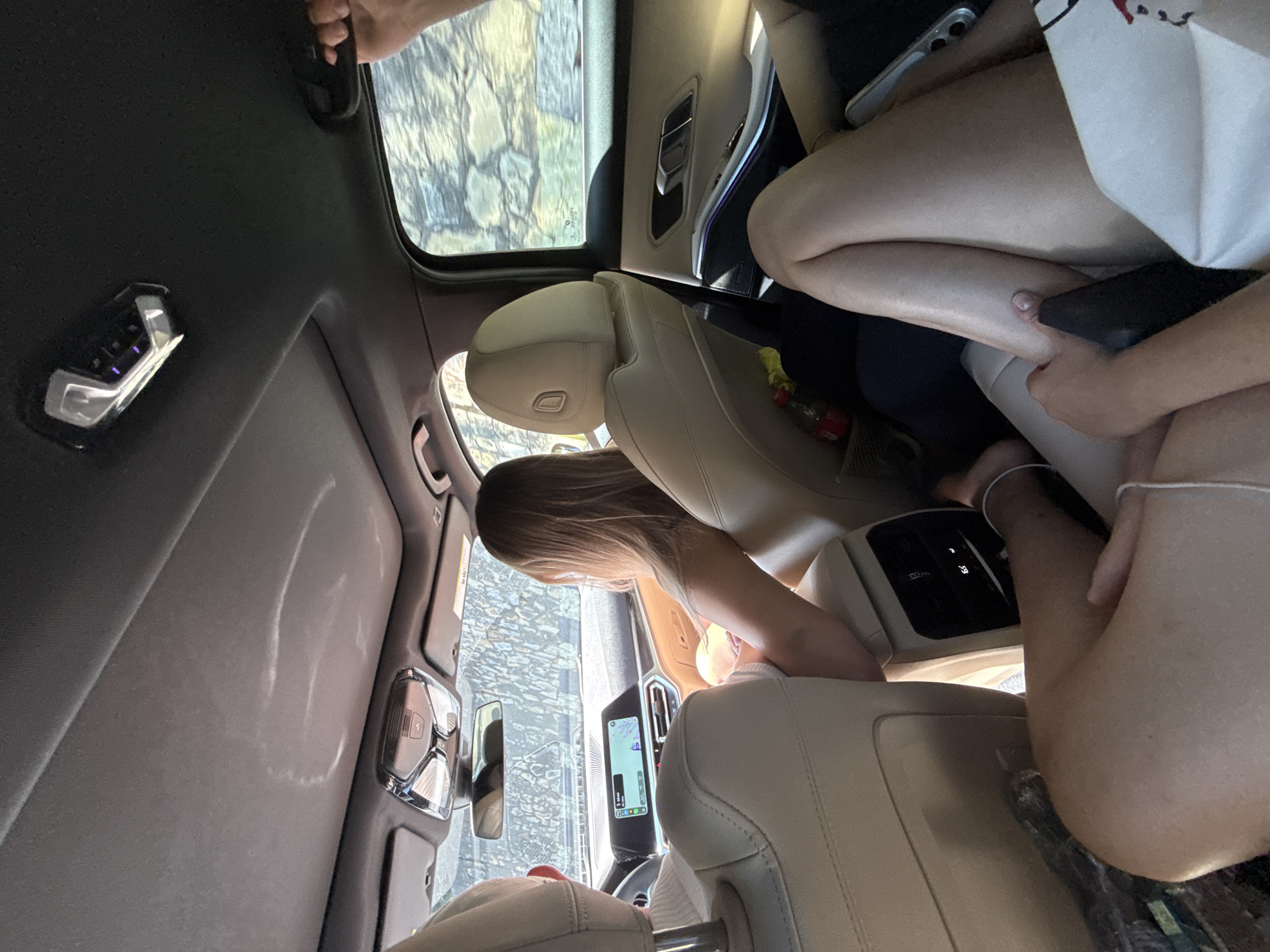 A group of people are inside a car on a paved road. The car's interior is visible, including the sunroof, dashboard, and seats. The passengers are mostly obscured, but one individual is visible in the passenger seat, with another in the back seat. The exterior of the car shows a rocky hillside and some trees. The image appears to have been taken during the daytime. The location is Oituz, Romania.