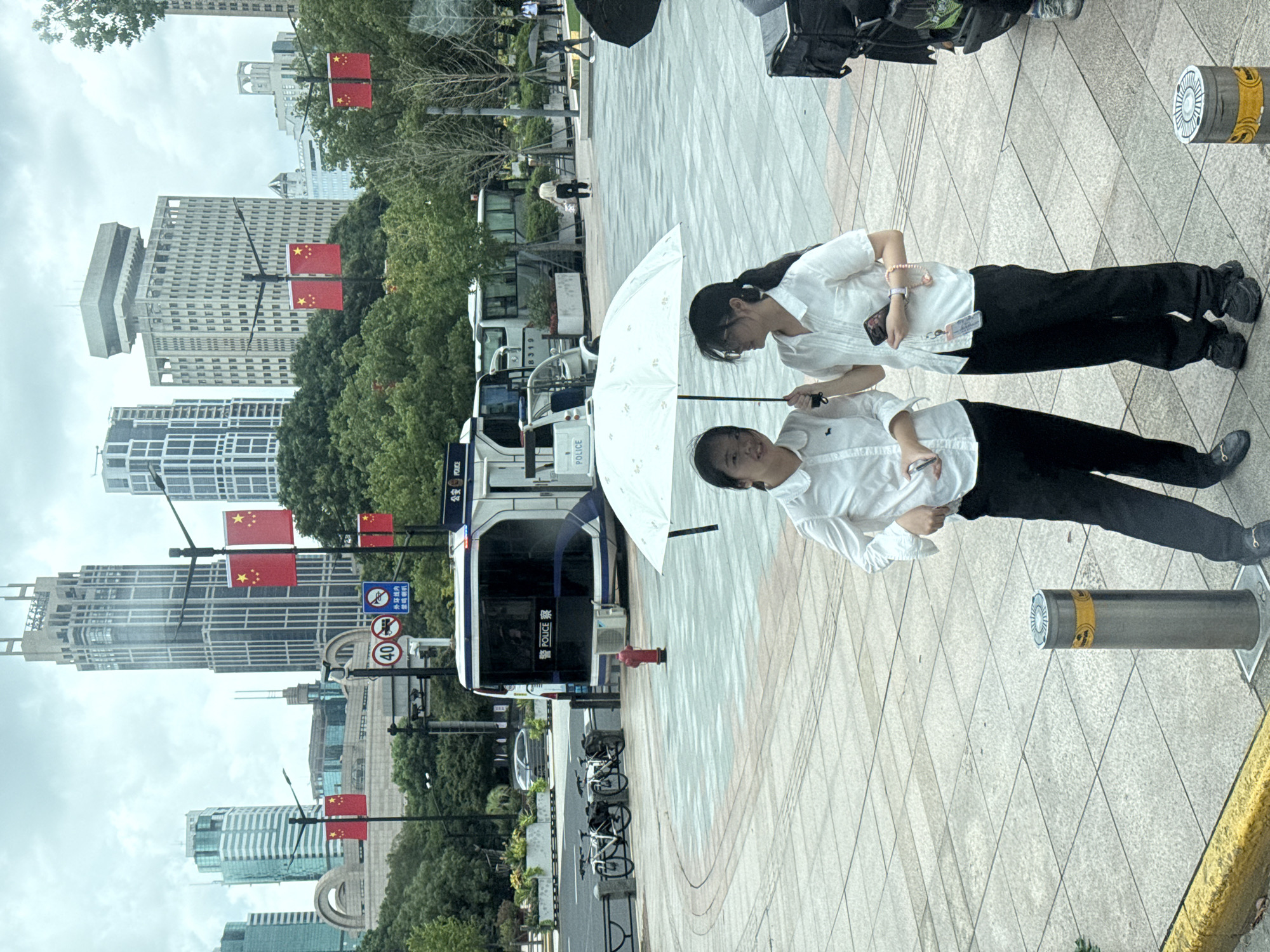 In Shanghai, China, on a cloudy day, two young women stand under a white umbrella on a paved plaza. Both are wearing white collared shirts and black pants. The woman on the left holds a phone in her left hand, while the woman on the right holds a phone and a pink bracelet on her left wrist. Behind them, a white police vehicle with blue accents is parked. Several bicycles are chained to a fence to the left of the police vehicle, and a red fire hydrant is visible nearby. In the background, modern skyscrapers and trees are seen under an overcast sky. Chinese flags are displayed on poles along the street. The overall scene suggests a public urban environment, possibly during a time of official presence or activity, given the police vehicle and flags.