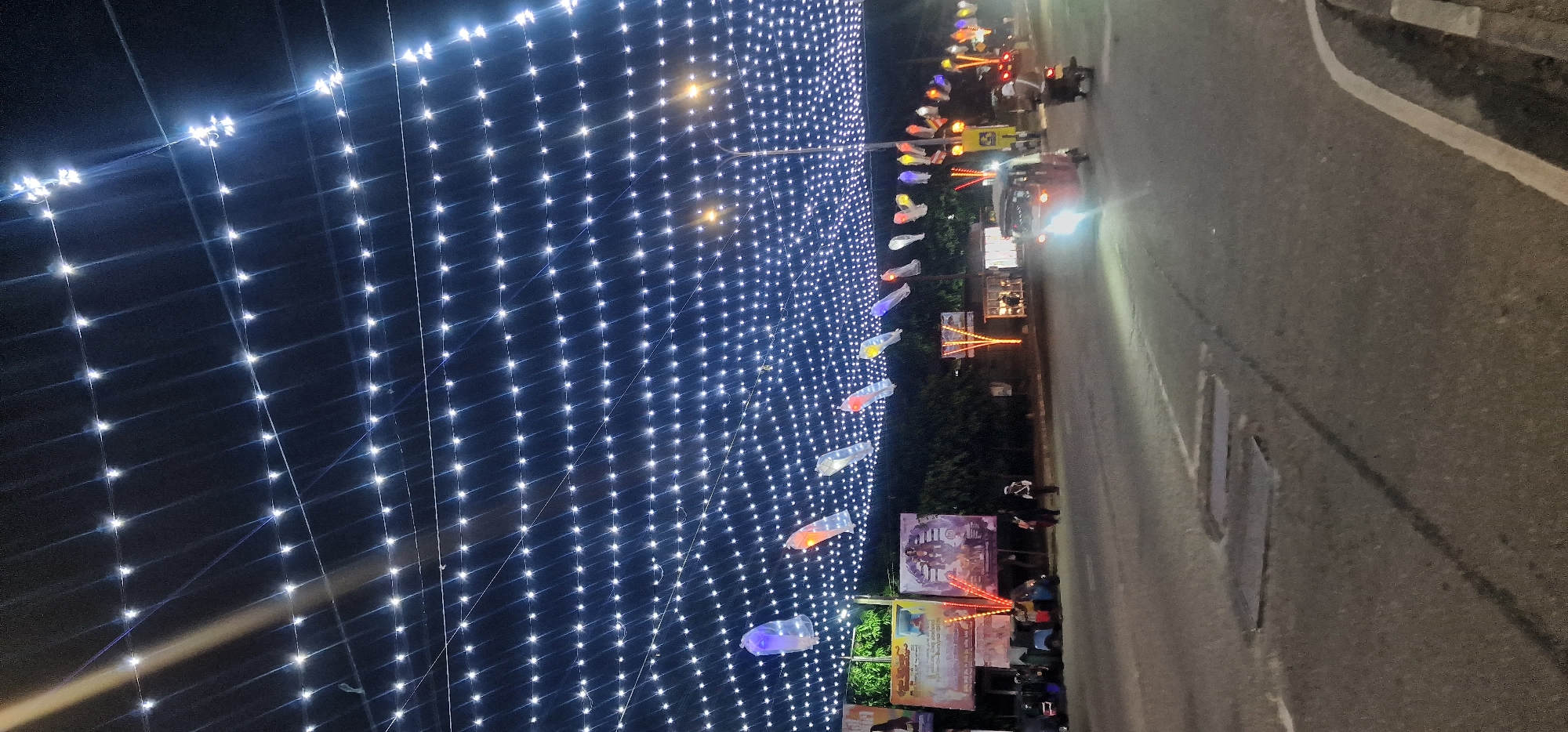 A nighttime scene in Matara, Sri Lanka, features a display of white lights stretching across the upper left of the frame. To the right, there is a road with a white line marking. Along the side of the road, there are various posters and banners visible, as well as some people and a couple of motorcycles. Decorative lanterns in colors of blue, purple, orange, and white hang above the road. The environment appears to be a street setting with lighting and decorations for an event or festival.