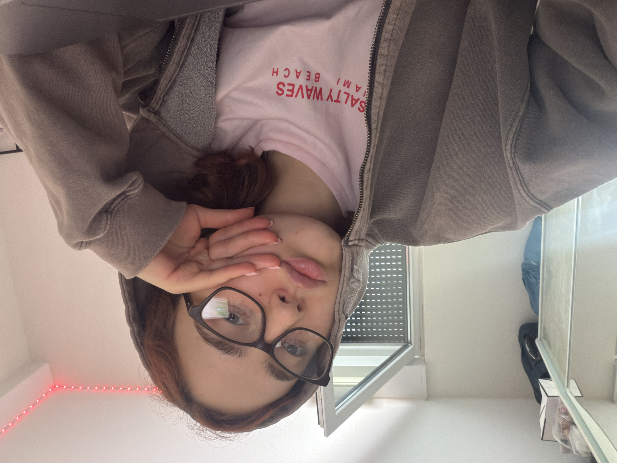 A young woman with reddish-brown hair and glasses rests her face on her hand, her mouth slightly pouted. She is wearing a light pink t-shirt with the text "SALTY WAVES MIAMI BEACH" in red letters, and a gray hooded sweatshirt. The setting appears to be indoors, possibly a bedroom or dorm room, with a window and a shelf visible in the background. Red string lights are visible along the top of the wall. There is no indication that this image was taken in Strasbourg, France.