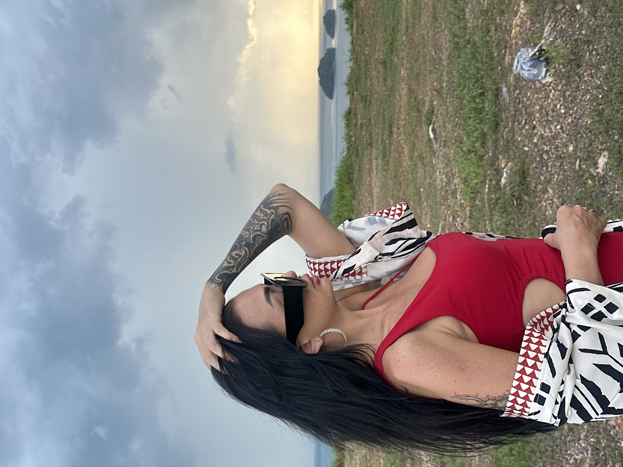 The image features a solo adult female standing in profile on an elevated, sparsely vegetated overlook in Baan Pang Ka, Thailand. She has long, dark hair, dark rectangular sunglasses, and large, ornate hoop earrings. Tattoos are visible on her left forearm and extending onto her upper left arm, as well as a smaller tattoo on her right upper arm. Her left hand is raised, fingers splayed, resting on the crown of her head. She wears a red sleeveless garment with a cut-out on the right side of her torso, covered partially by a draped long-sleeved shawl featuring a black, white, and red geometric pattern. Her gaze is directed towards the upper right, away from the camera, across a calm body of water (sea) dotted with three dark, small, vegetated islands in the mid-distance. The sky above is heavily overcast with grey clouds on the left, transitioning to a brighter, golden-yellow light on the right horizon, suggesting either dawn or dusk. The foreground consists of dry, patchy earth with scattered green and brown grass and small rocks. A small, light-colored object, possibly debris, lies on the ground in the lower right corner.