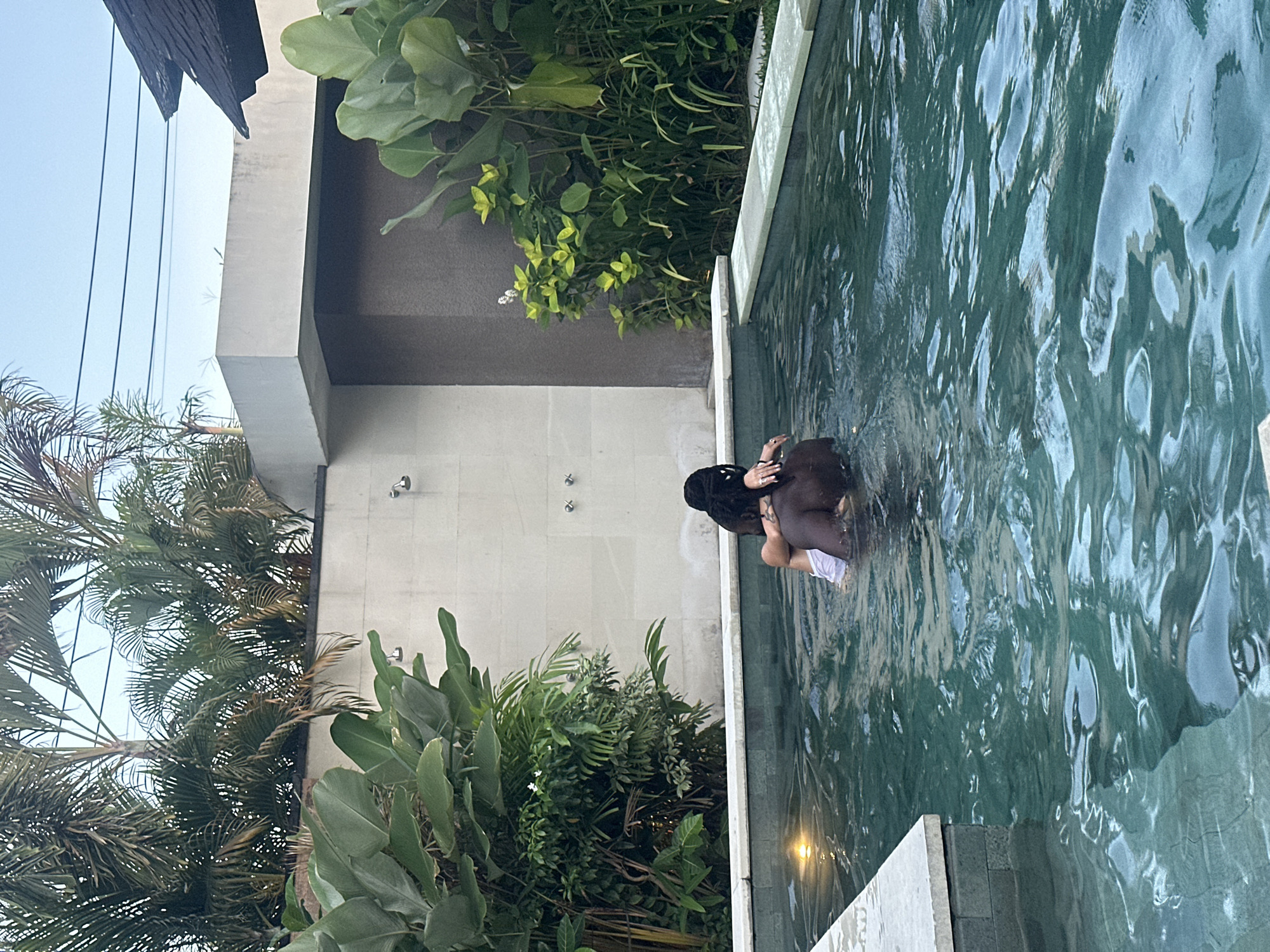 The image, taken in what appears to be North Kuta, Indonesia, shows a partial outdoor view of what seems to be a building with vegetation, possibly a hotel or resort, and a pool. A person with dark hair is in the pool, partially submerged. There is lush tropical greenery surrounding the pool and near the building. There are a few light fixtures visible near the pool's edge.