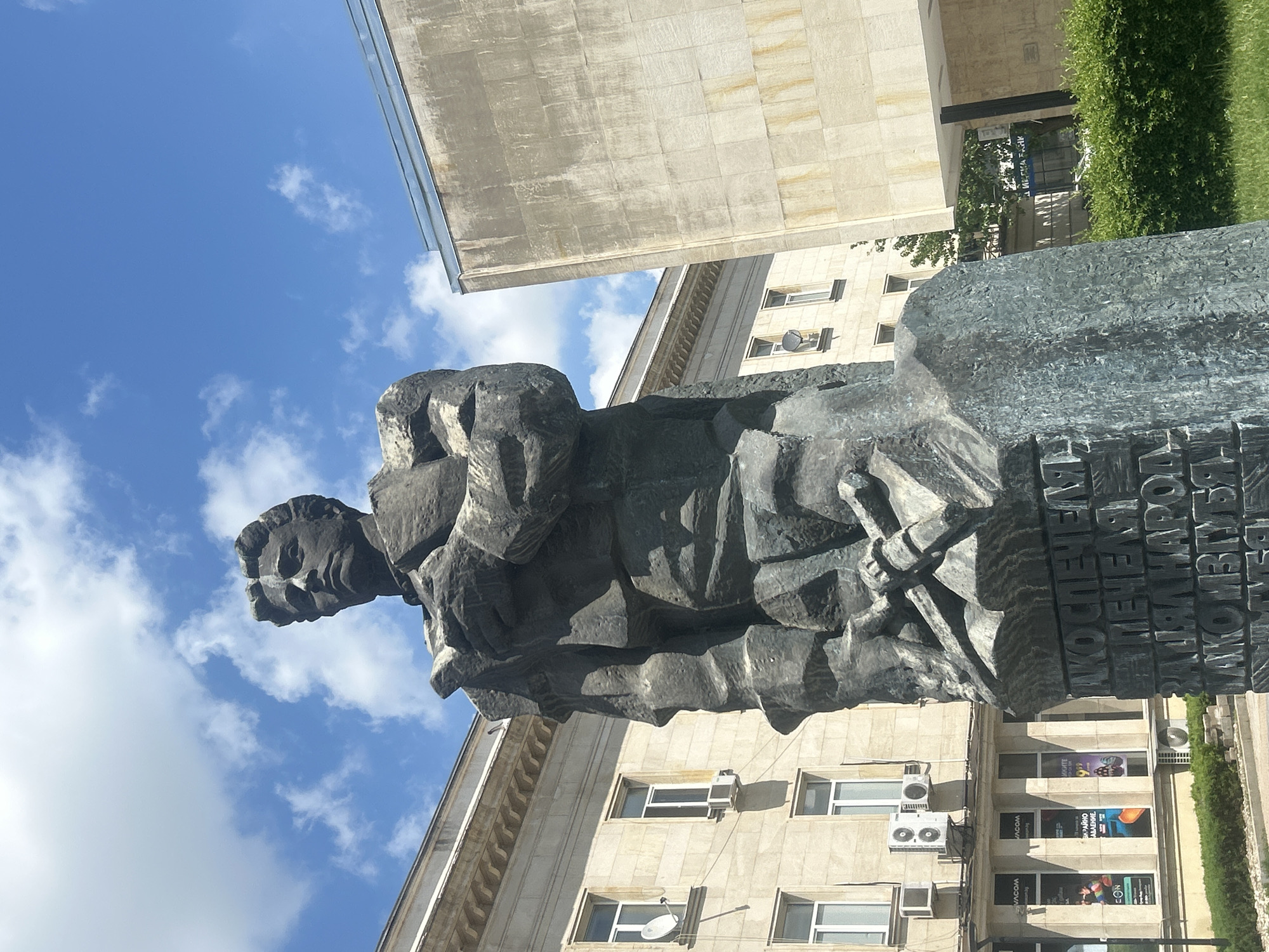 The image depicts a monumental stone statue of a man, positioned in an urban environment against a backdrop of buildings and a partly cloudy sky. The statue features a rugged, textured surface, and the lower part includes inscribed text, likely in Cyrillic. A stone building with a lighter facade features windows, satellite dishes, and air conditioning units. A modern building with advertisements appears in the background. To the right of the statue, there is a strip of green foliage. The statue is located in Pleven, Bulgaria.