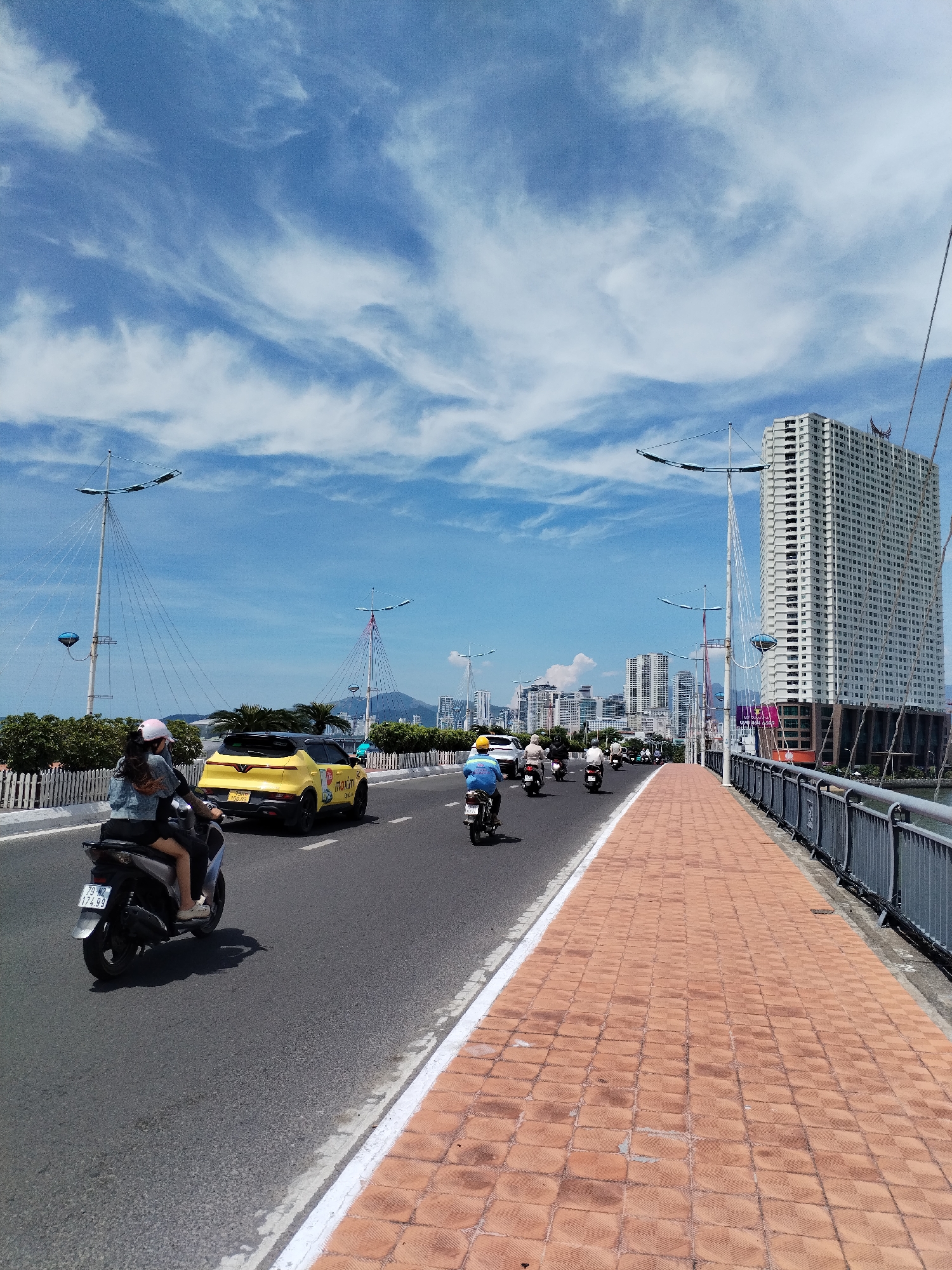 This image captures a view of the Dragon Bridge in Da Nang, Vietnam, on a clear, partly cloudy day. The bridge, spanning a body of water, features a pedestrian walkway paved with orange bricks on the right side, separated from the road by a metal railing. The road itself is asphalt and carries several vehicles, including a bright yellow car with "Maxim" branding and multiple motorcycles. On the left side of the road, two individuals are riding a motorcycle, with the passenger wearing a pink helmet and denim jacket. In the background, a large, modern high-rise building dominates the skyline, characteristic of a coastal city. The sky is a vibrant blue with wispy white clouds. The overall scene depicts a bustling urban environment with a focus on transportation and modern architecture.