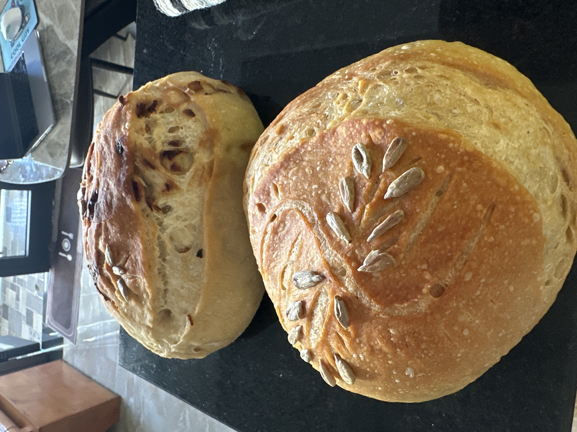 Two loaves of bread rest on a dark, possibly granite, countertop. The loaf on the right is round and decorated with sunflower seeds arranged in a stem-like pattern. It has a golden-brown crust. The loaf on the left is more oblong, with a lighter crust and embedded seeds, possibly onions, charred on the surface. The background is slightly out of focus, but includes a tiled wall, a blurred object labeled "Cool Guy", and other kitchen items. There is no visual indication of the location being Bac Nha Trang Ward, Vietnam.
