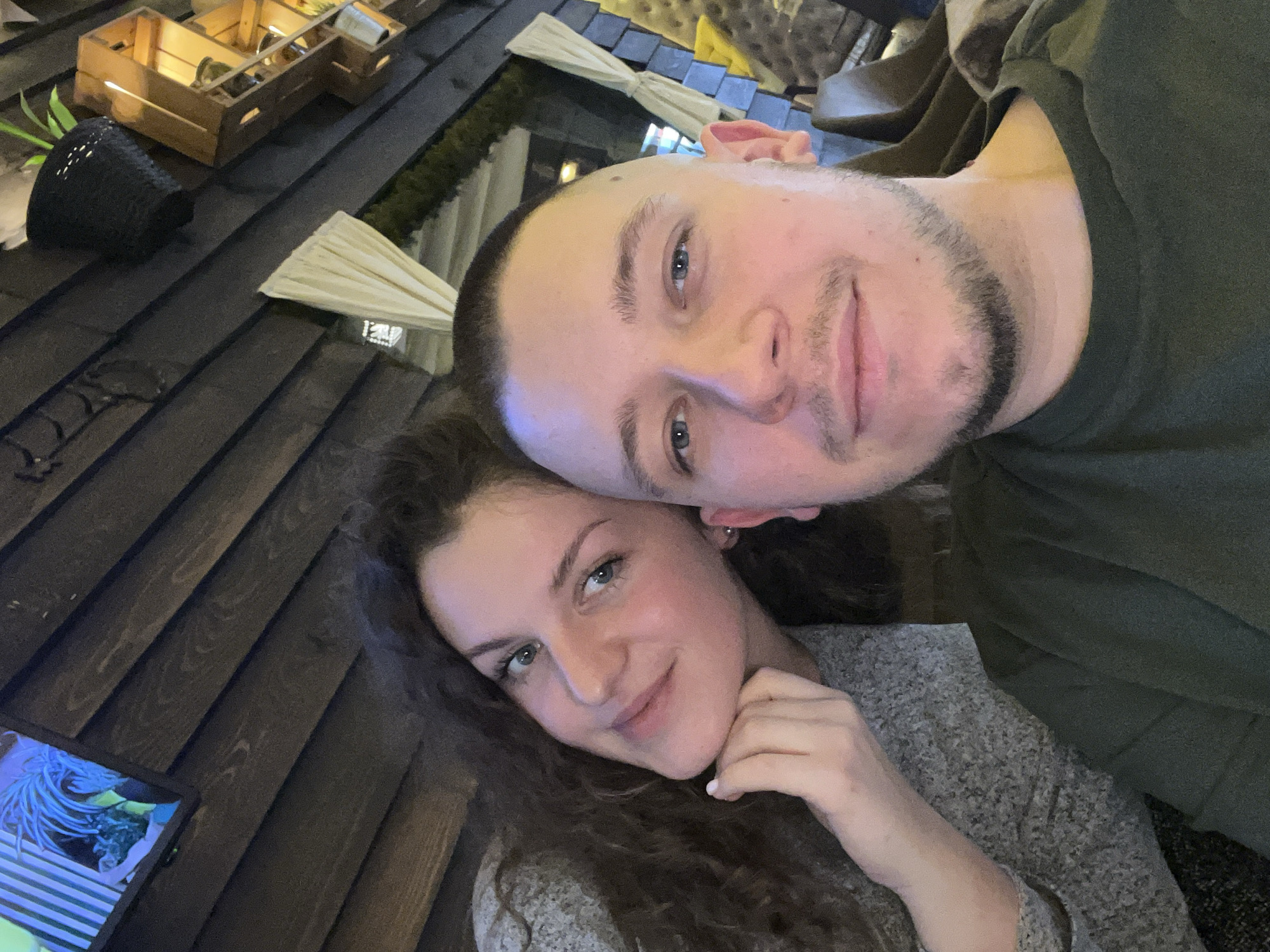 Captured at an undisclosed location in Sofia, Bulgaria, the image presents a selfie featuring a Caucasian man and woman. The man, positioned slightly higher, sports a close-cropped haircut, light eyes, and a trimmed beard. He wears a dark green shirt. The woman, situated below him, has long, wavy brown hair and light eyes; she's wearing a gray sweater and rests her chin on her hand. The background shows wooden paneling, what appears to be a table with decorative items like a wooden crate containing objects and a black woven pot, and possible outdoor lighting or decorations. A screen or device is visible in the lower left corner. The subjects' expressions suggest a relaxed and intimate moment.