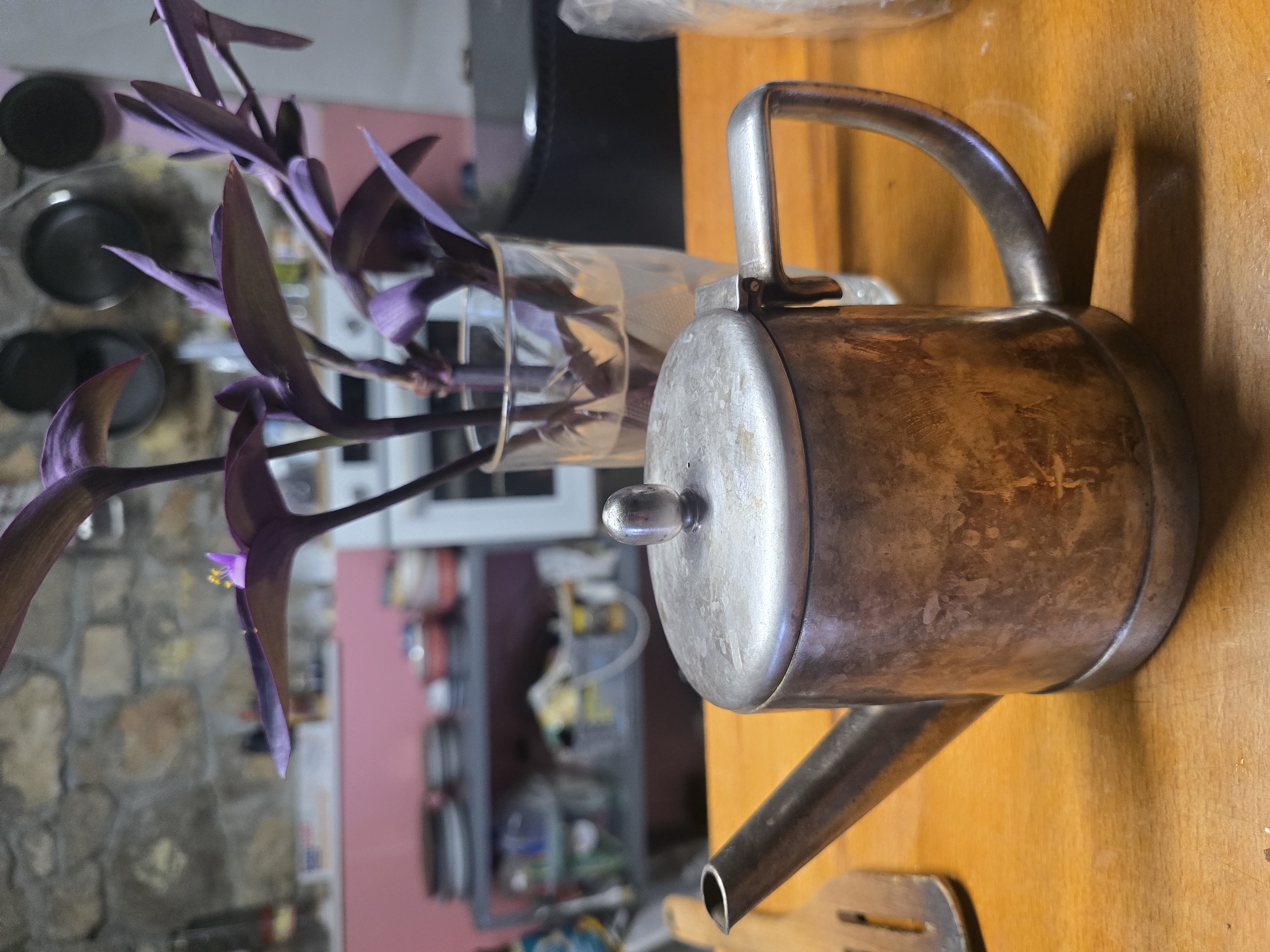 A close-up, eye-level, medium shot captures a silver teapot on a wooden surface, with a blurry background of a kitchen. The teapot is in the foreground, to the right of the center, and is oriented at a slight angle with its spout pointing left. The teapot is made of metal with visible patina and scratches, suggesting age and use. It has a rounded body, a lid with a small knob on top, a C-shaped handle on the right side, and a narrow spout on the left. To the left of the teapot, a portion of a wooden utensil with a slotted surface is visible. Behind the teapot, a glass of water holds a plant with broad, purple leaves and a small yellow flower. The background is out of focus, revealing shelves with various items, including what appear to be dishes, containers, and cooking utensils. The wall behind the shelves is made of rough, grey stone. The overall lighting is dim, casting shadows on the wooden surface.