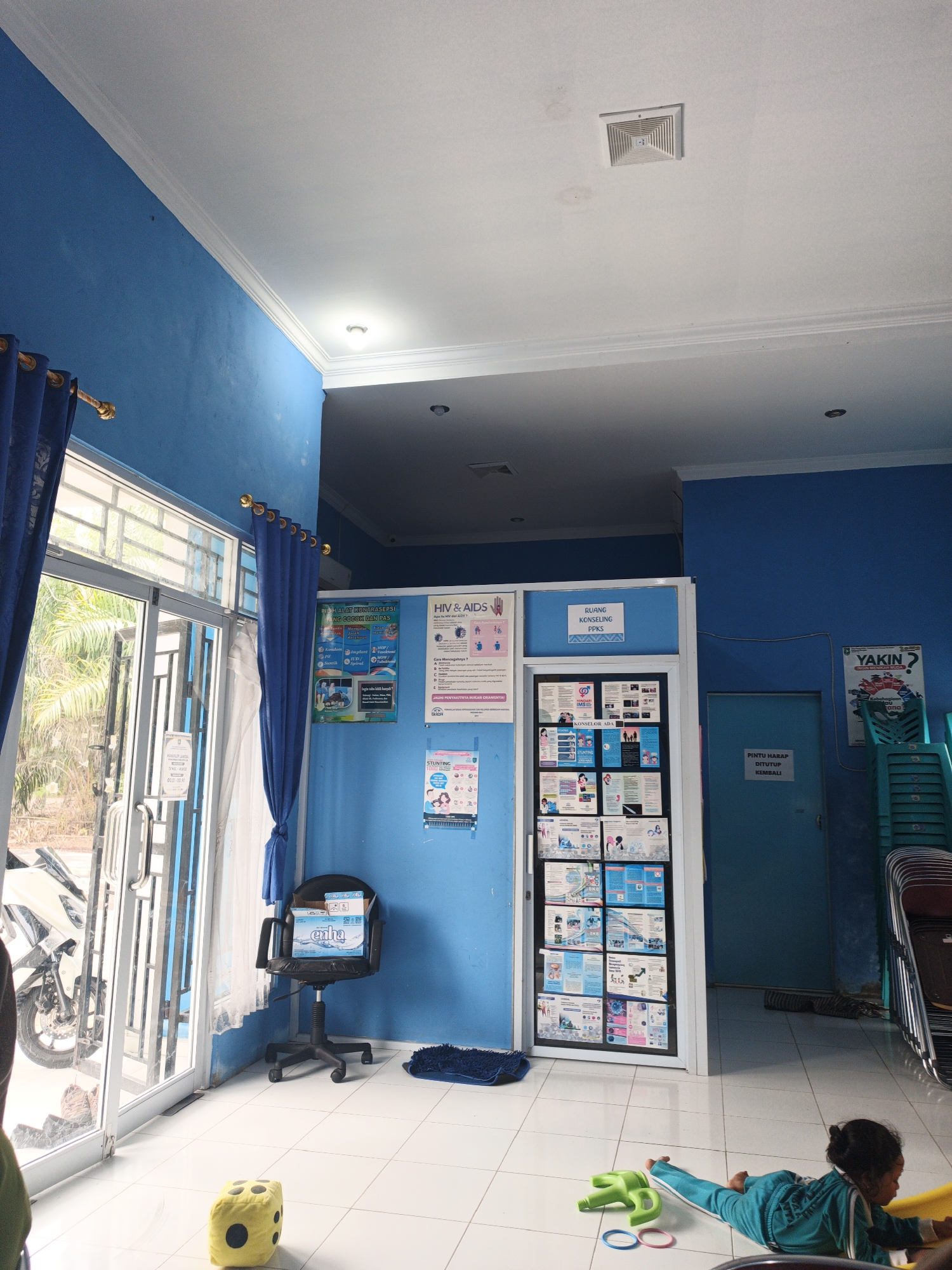 The image depicts the interior of a room, possibly a clinic or community health center, painted in vibrant blue. A glass door with metal accents is visible on the left, revealing a glimpse of the outdoors with greenery and a motorcycle parked outside. Blue curtains hang to the sides of the door. Inside, a white-tiled floor spans the space.

To the right of the door, a black office chair with wheels is positioned. On the chair sits a blue and white box of "exha" products. Near the chair, a yellow dice-shaped toy rests on the floor. A dark blue rug is placed further into the room.

A large display board is mounted on the wall, showcasing various informational posters and pamphlets. Above this display, a sign reads "RUANG KONSELING PPKS". Another poster on the wall to the right has the text "HIV & AIDS" and information about contraception. Further to the right, a teal-colored door is present, and stacked chairs are visible behind it. A poster with the word "YAKIN?" is also on the right wall.

In the foreground, a young child, dressed in a teal outfit, is lying on the floor, seemingly playing with colorful toys. The ceiling is white, with some visible light fixtures and a ventilation grate. The overall atmosphere appears to be a functional space with an emphasis on health information and patient services. The location is identified as Resam Lapis, Indonesia.