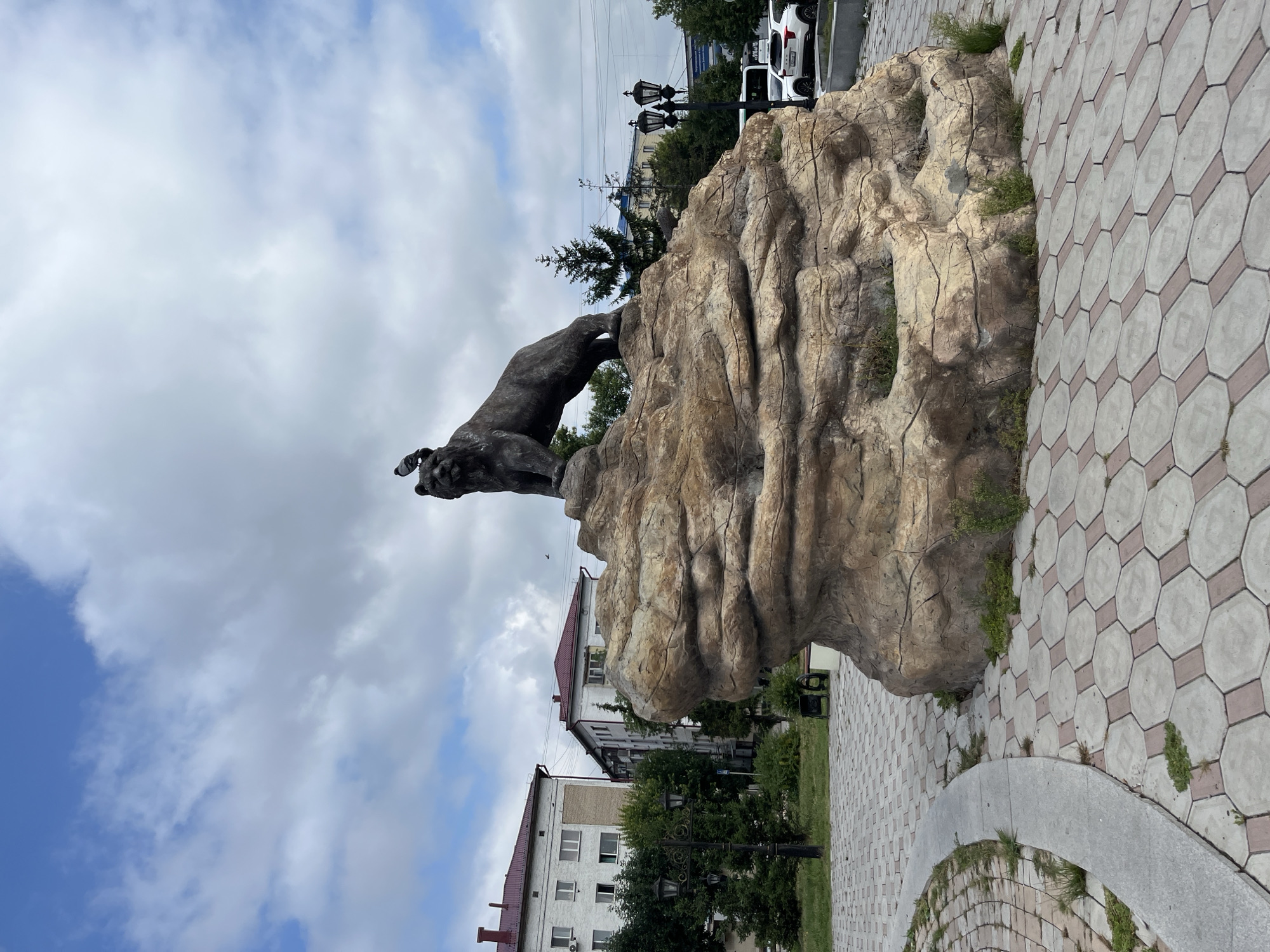 This is a medium shot, eye-level, outdoor, full-color photograph of a statue of a bear on a rock formation in the city of Petropavl, Kazakhstan. The statue is made of dark metal and depicts a bear in a standing pose, with one paw raised. A pigeon is perched on the bear's head. The rock formation is large and textured, with areas of light brown and tan. It is situated on a paved area with a curved walkway and octagonal grey and pink paving stones. In the background, there are several multi-story apartment buildings with white and red facades, green trees, and street lamps. A street with parked cars and a white bus is visible in the distance. The sky is cloudy with patches of blue. This statue is a prominent landmark in Petropavl, likely a symbol of the city or region.