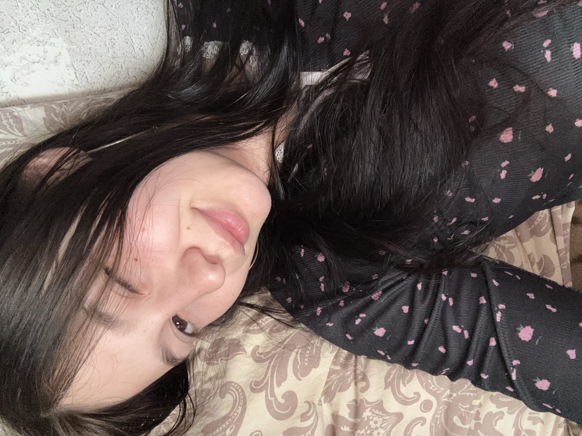 A young female with long, dark hair is depicted lying down in an indoor setting in Фоки, Russia. She is winking with her right eye and has a subtle smile. Her left eye is open, displaying a dark iris. The subject has light skin with visible small facial moles. She is wearing a dark, possibly ribbed, long-sleeved garment patterned with small, irregular pink shapes. A thin, light-colored hoop earring is visible on her left ear. She rests on a pillow or bedsheet with a light brown paisley-like pattern. A light-colored, textured wall is visible in the background to the right of her head.