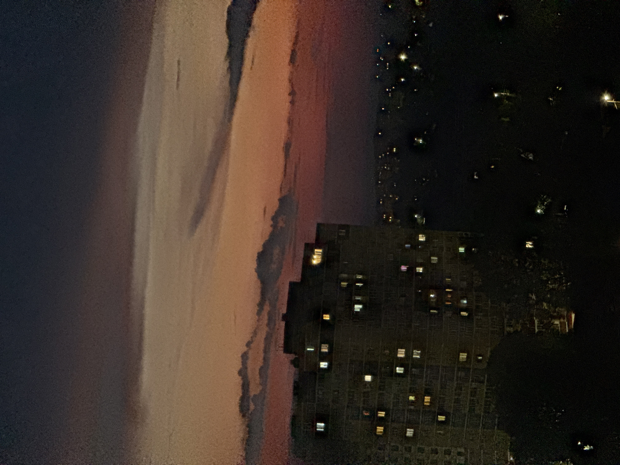 An aerial nighttime view captures a residential apartment building against a dusky sky in Izhevsk, Russia. The sky displays a gradient of deep blues and purples at the top, transitioning to lighter oranges and reds closer to the horizon, suggesting twilight or sunset. Clouds are visible, with darker, denser formations obscuring some of the lighter sky. The apartment building, a large, multi-story structure, is illuminated by numerous lit windows, scattered across its facade. In the foreground and extending into the background, the cityscape is visible as a dark silhouette, punctuated by scattered streetlights and other building lights, indicating a populated urban area. Trees or other vegetation are present in the lower foreground, appearing as dark masses. The overall scene depicts a residential area at dusk or night.