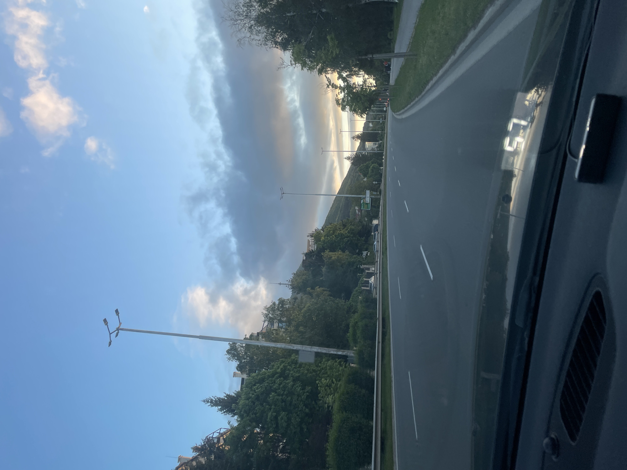A roadside scene is presented in this vertically-oriented photograph taken from inside a vehicle. A portion of the dashboard is visible on the right side. Outside the car, a straight road stretches into the distance, flanked by green trees and foliage. On the left side of the road, street lights stand tall against a backdrop of a blue sky with scattered clouds. Some buildings can be seen nestled among the trees. There is a mountain in the distance. The city location is not identified.