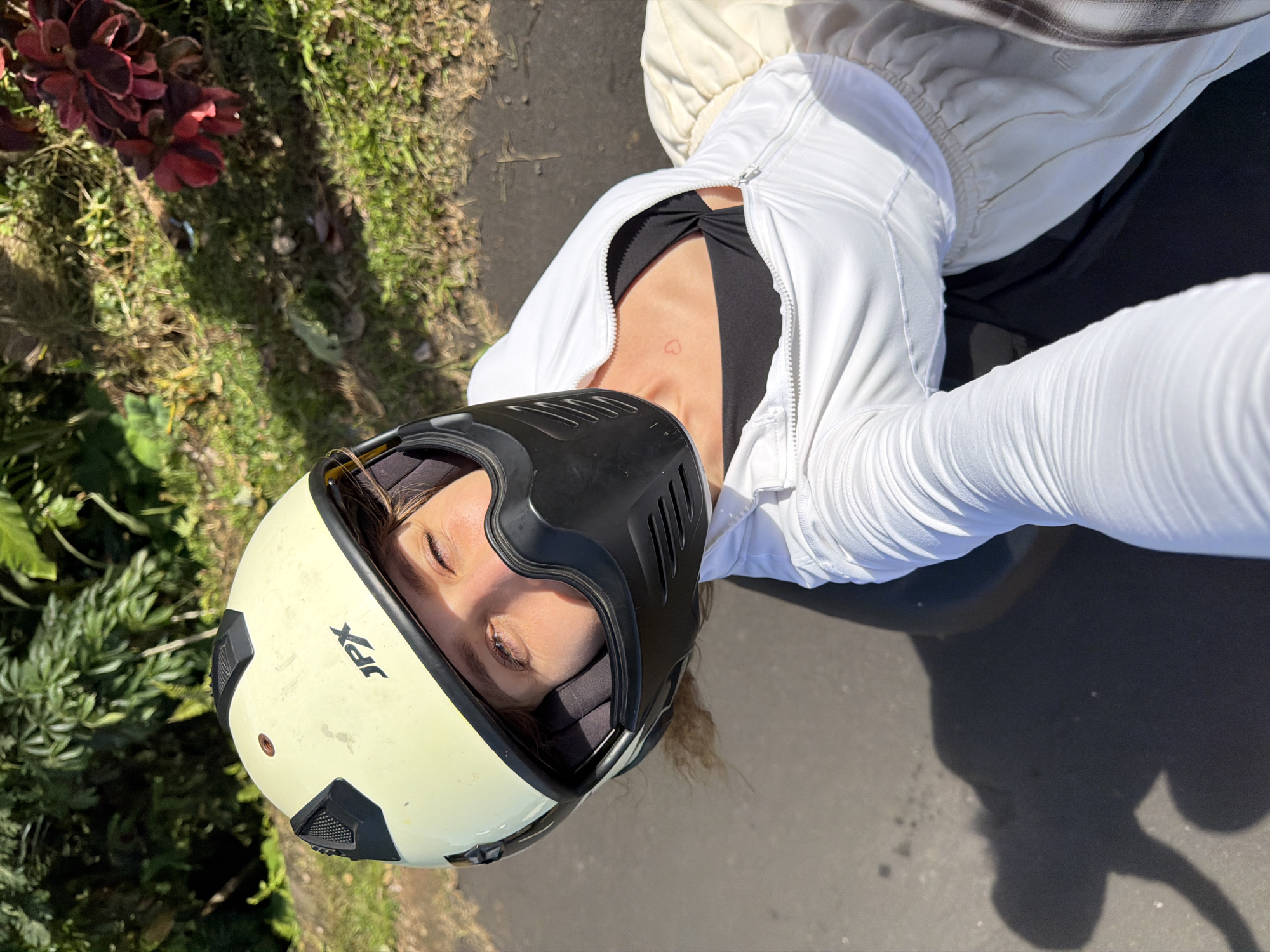 A high-angle, selfie-style shot captures a woman from the chest up, wearing a light yellow motorcycle helmet with a black face mask and white zip-up jacket. She is looking at the camera with her eyes partially closed. A faint red heart tattoo is visible on her upper chest, above a black sports bra. The background consists of lush green foliage and grass, with a patch of red-leafed plants on the right. The ground appears to be asphalt, with a shadow of the woman and her motorcycle visible on the left. The lighting suggests it is daytime with bright sunlight. The scene is located in Penebel, Indonesia, likely in a rural or suburban area given the natural surroundings.