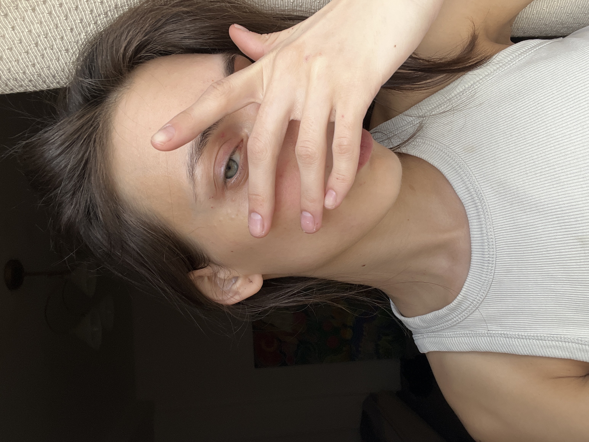 This media file presents a close-up, angled portrait of a person, likely female, indoors in Krasnoyarsk, Russia. The subject has fair skin, dark brown hair, and a striking green eye that is clearly visible. They are wearing a light-colored, ribbed tank top, appearing either white or light grey.
The person is depicted in a casual pose, with their right hand partially covering their face. They are looking towards the camera through the fingers of their hand, allowing one eye to be seen. Their nails are natural, showing faint white tips, and appear unpolished. Minor skin details, such as small blemishes or moles, are visible on the forehead, the back of the hand, and the neck/shoulder area. The dark brown hair appears natural and slightly disheveled, framing the face.
The setting is indoors, with a textured, light-colored fabric—possibly a couch or bed headboard—visible in the upper left background. The lower right background is dark and indistinct, suggesting a dimly lit room, though a subtle, colorful, abstract pattern or painting can be discerned within the shadows. The lighting is soft and consistent with indoor conditions, not indicating bright outdoor illumination.
The activity captured is posing for a photograph. The expression conveyed through the visible eye and the partially obscured face suggests a calm, possibly introspective or coy mood. There is no visible text present in the image.