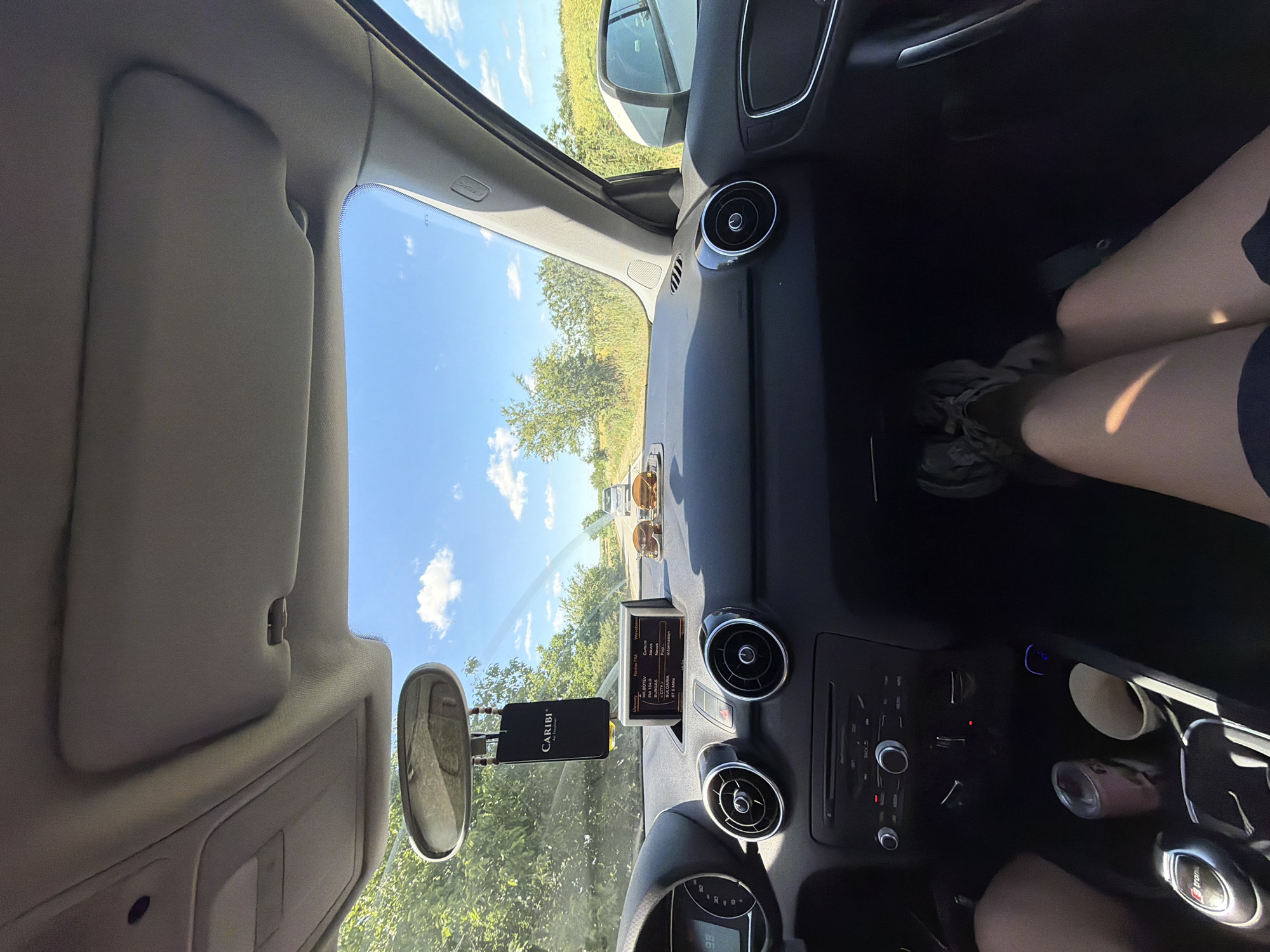 The image is taken from the interior of a car, looking forward through the windshield on a sunny day. The dashboard is visible, including the car's radio display which shows station information. A black air freshener hangs from the rearview mirror. On the dashboard, there are two sunglasses and two hamburger figurines. Outside, the road stretches ahead, lined with trees and greenery. Further down the road, two other cars are visible. The sky is blue with scattered white clouds. The passenger's feet are visible in the foreground, wearing sneakers. The scene appears to be a road trip. The location is identified as being in the city of Resen, Bulgaria.