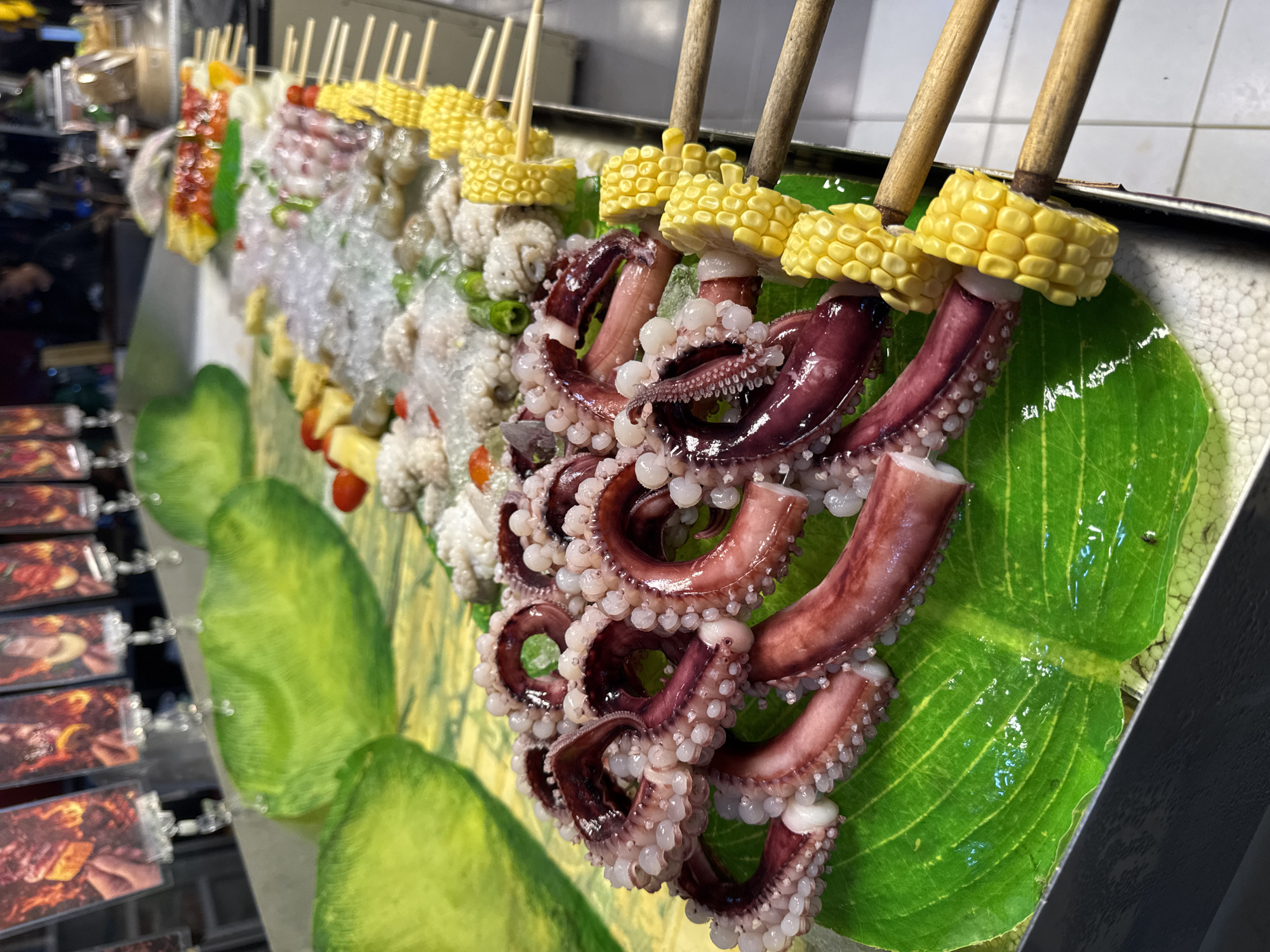 At a food stall in Karon, Thailand, a display of food items on skewers is presented on a metal counter. In the foreground, several large octopus tentacles, their suckers prominent, are arranged on a bright green, glossy leaf. To the right of the octopus, skewers of corn on the cob are visible. Further back, a variety of other ingredients, including what appears to be shrimp, small octopuses, and some vegetables, are displayed on skewers, some covered in crushed ice. The background shows more food items and indistinct figures of people, suggesting a busy market or street food environment.
