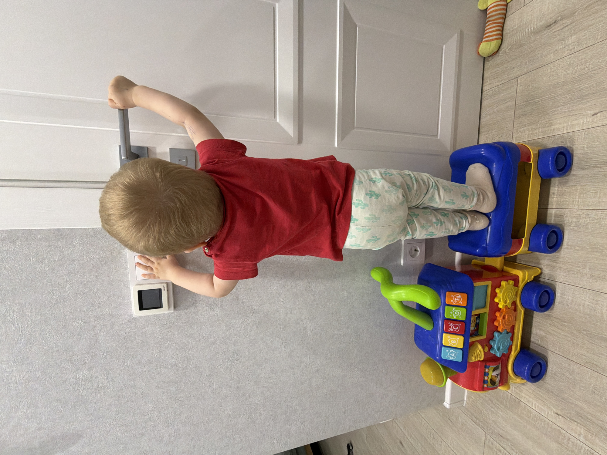 A small child with blonde hair and fair skin, wearing a red shirt and light-colored pants with a pattern of small green cacti, is standing in front of a closed white door with raised panel details. The child's left hand is on the door handle and their right hand is touching a wall-mounted thermostat. The child appears to be standing on the back of a multi-colored toy walker.
The door and thermostat are mounted on a light grey wall. Part of a striped sock is visible near the top of the door. The floor appears to be laminate flooring with a light wood grain pattern.
There is no evidence to suggest that the photograph was taken in Votkinsk, Russia.