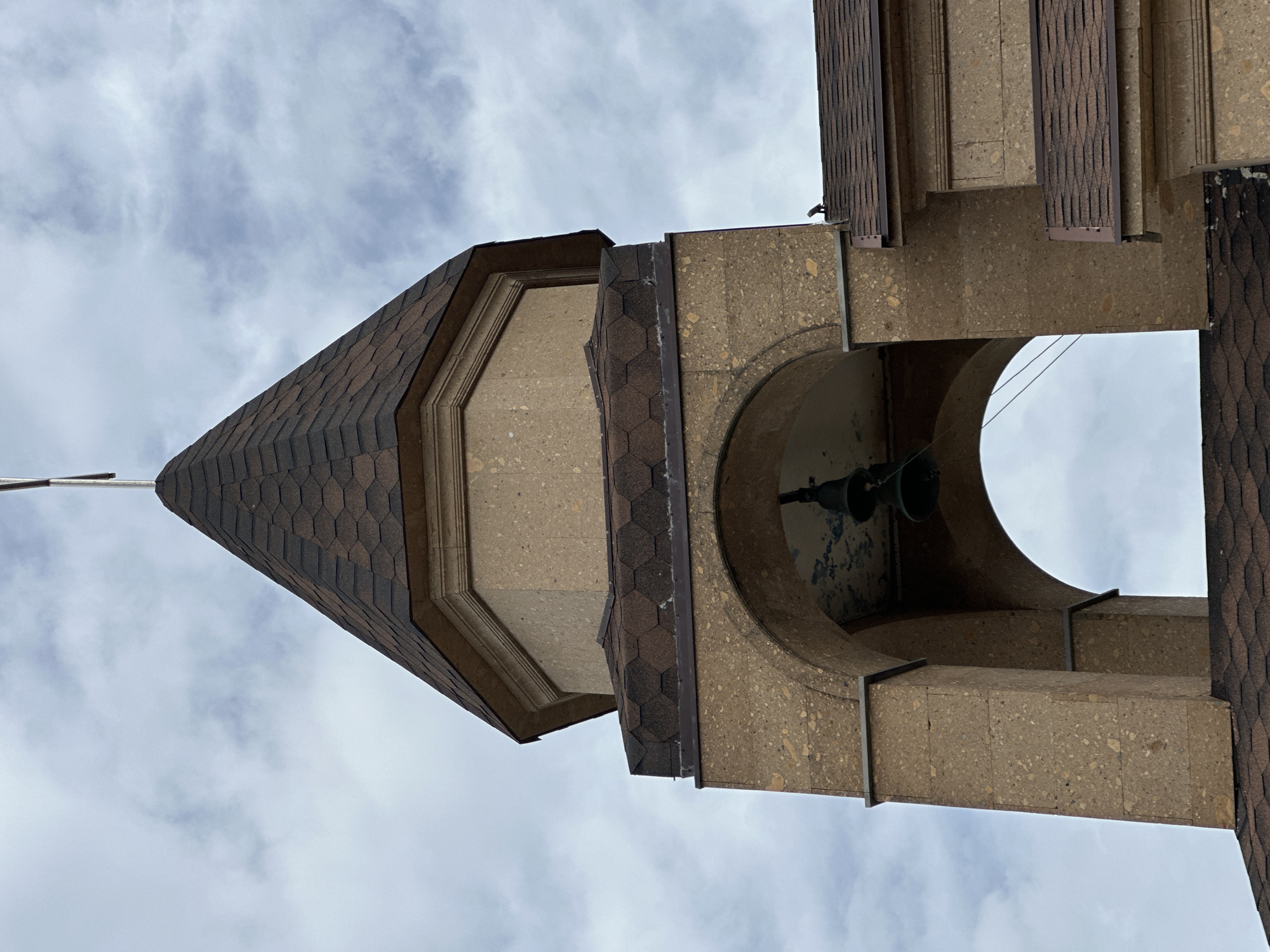 View from below of the steeple of a building, possibly a church. The steeple has a conical roof with brown hexagonal shingles. The tower is constructed of a tan stone with an arched opening containing a bell. The background is a cloudy sky. The location in the city of Novomikhaylovsky, Russia is not possible to infer based on the content of this image.