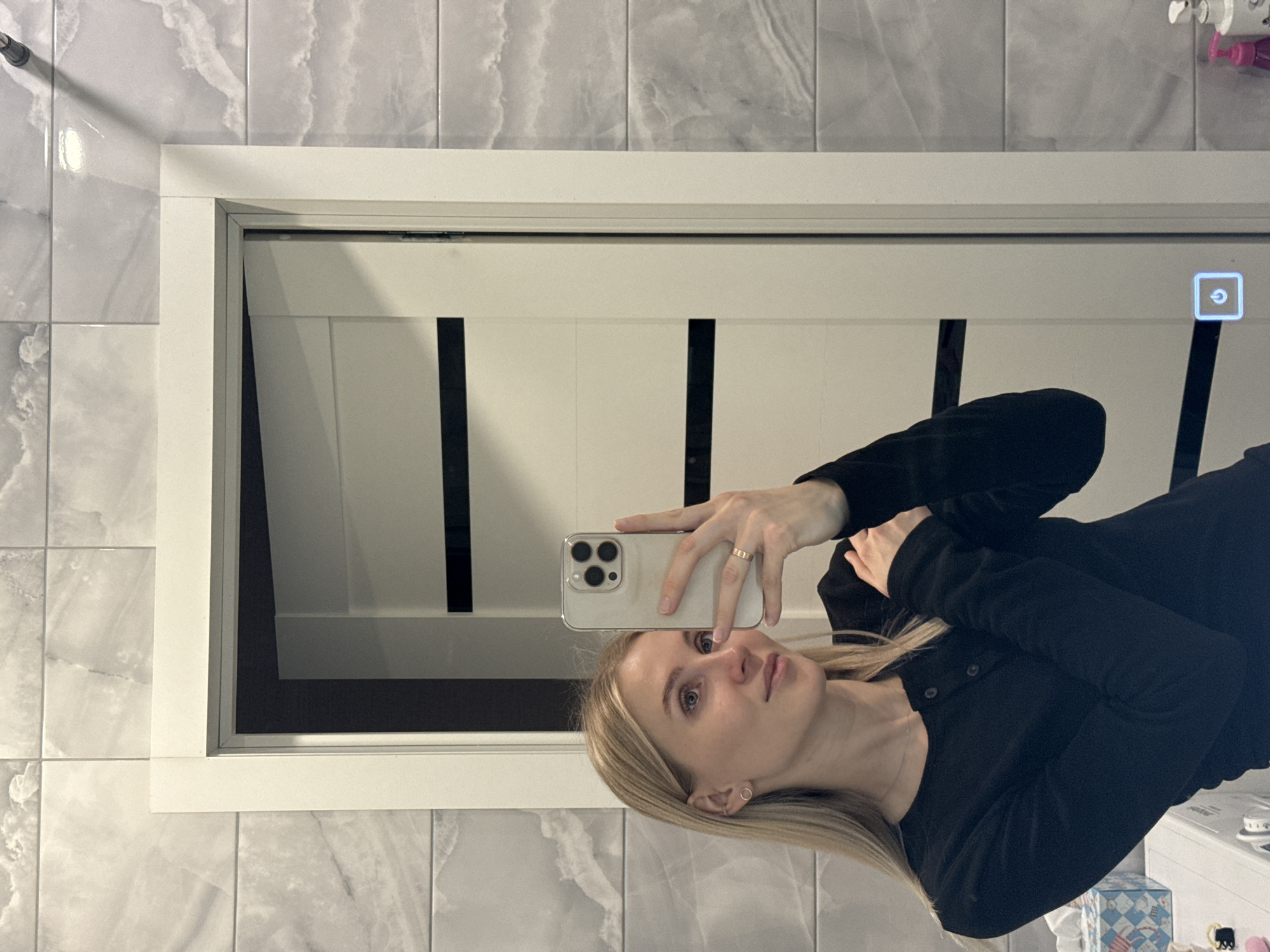A woman with blond hair is taking a selfie in a bathroom. She is wearing a black long-sleeved shirt. She is holding a smartphone in her right hand, taking the picture in a mirror. The bathroom has grey marble-like tiles on the walls and a white-framed mirror/cabinet with a white door that has black accents. Several toiletries are visible on a shelf above her head. Based on the environment, the location cannot be specifically determined as Izhevsk.