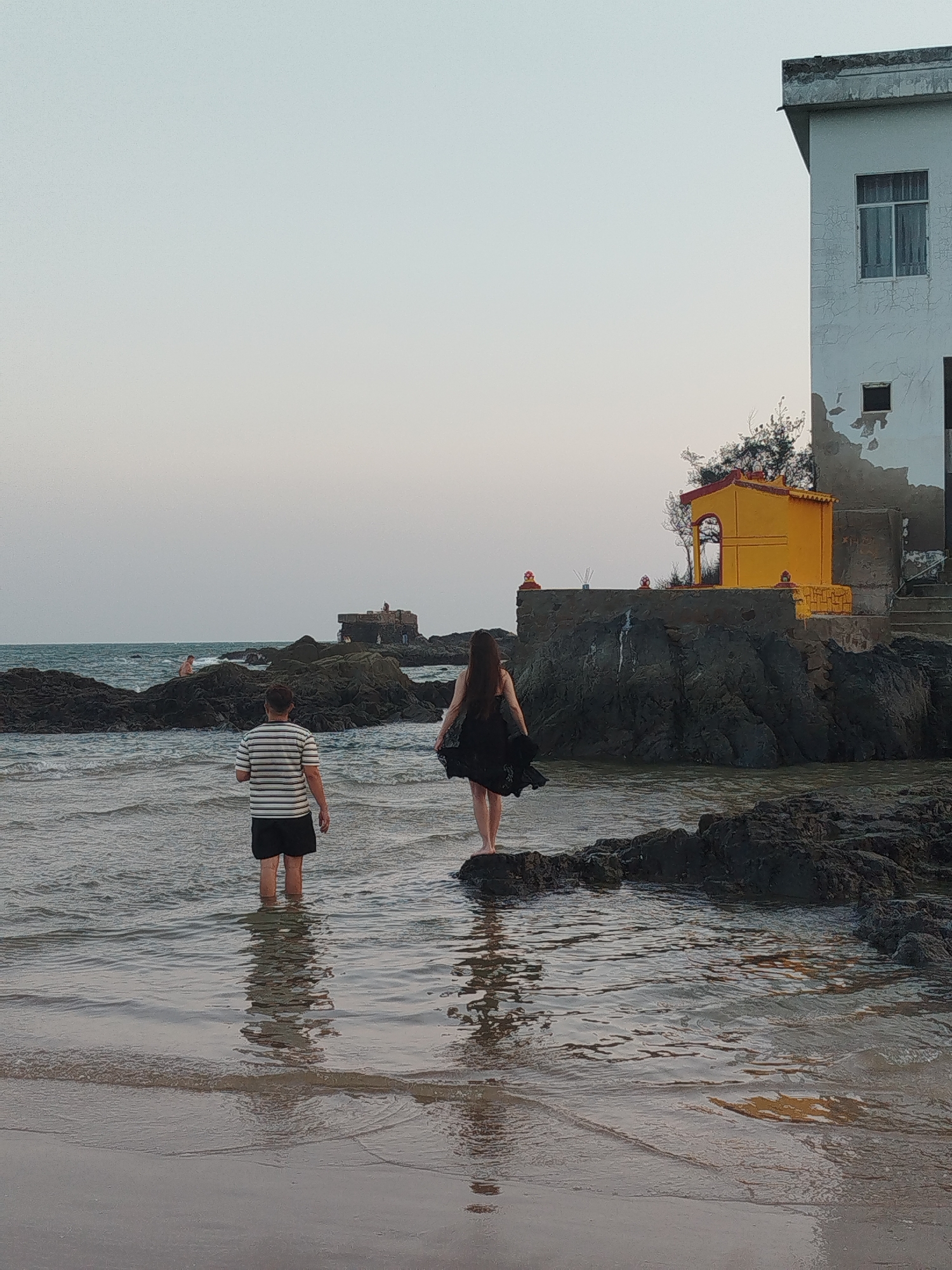 Two people, a man and a woman, are wading into the water at a beach. The man is wearing a striped shirt and black shorts, and is facing away from the camera. The woman is wearing a black dress and is also facing away from the camera.
Behind them is a rocky outcrop with a small yellow building on top. There is a larger, white building to the right of the yellow building. There are also other rocks and outcrops visible in the water, and a person can be seen swimming in the distance.
The beach is located in Ho Chi Minh City, Vietnam. The photograph was taken during the daytime.