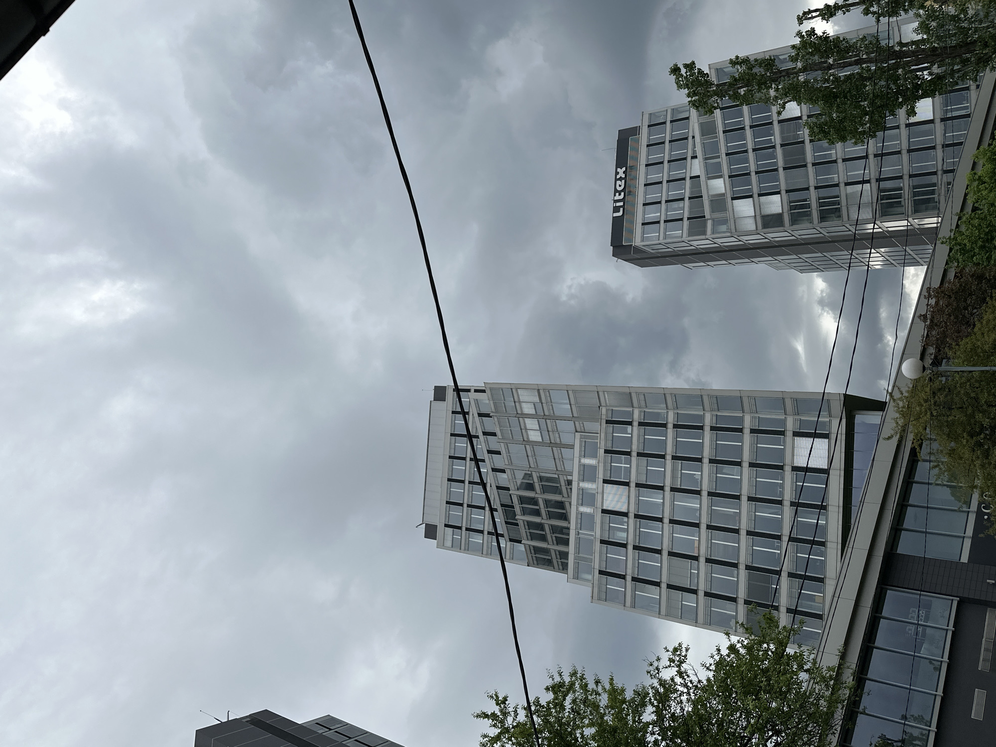 The image shows two high-rise buildings in Sofia, Bulgaria, under an overcast sky. The building on the right features "Litex" signage on its facade. Both buildings have a grid-like pattern of windows. Overhead power lines cut across the view. Some trees are visible at the bottom of the image.