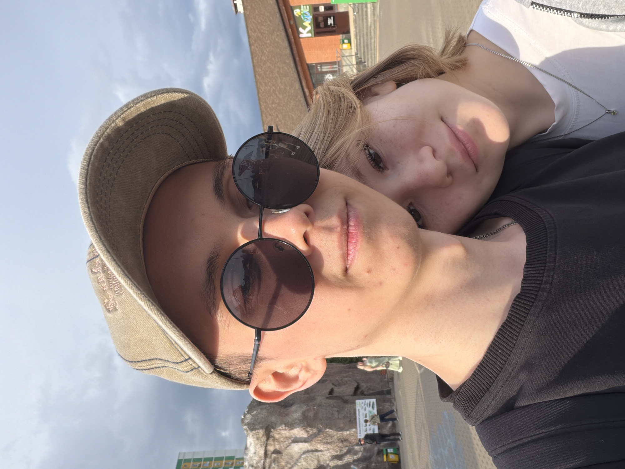 The image is a close-up selfie of two people. The person in the foreground is wearing a tan baseball cap and sunglasses. Their face is turned towards the camera, and their mouth is slightly open. The second person is behind them, resting their head on the first person's shoulder. They have blonde hair and are looking towards the camera with their eyes open.
The setting appears to be outdoors, possibly in a park or recreational area, with a cloudy sky in the background. In the distance, there is a building with signage, including the word "KARE" visible in larger font, and what looks like a zoo or animal enclosure with rock formations and people observing exhibits.
The time of day seems to be daylight, given the bright lighting and shadows. The mood appears casual and relaxed, possibly indicating a fun outing or a moment of shared affection. There are no explicit emotional cues beyond general contentment.
The image is a vertical orientation. The quality is typical of a smartphone photo.